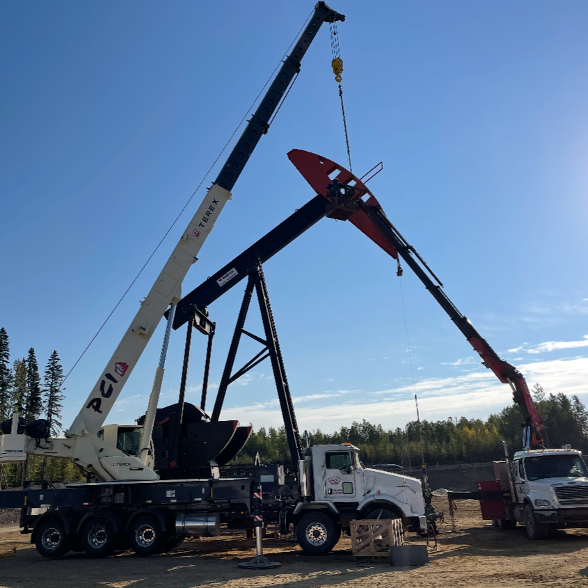 Two large cranes lifting and positioning a piece of construction equipment on a construction site with trees and a blue sky in the background.