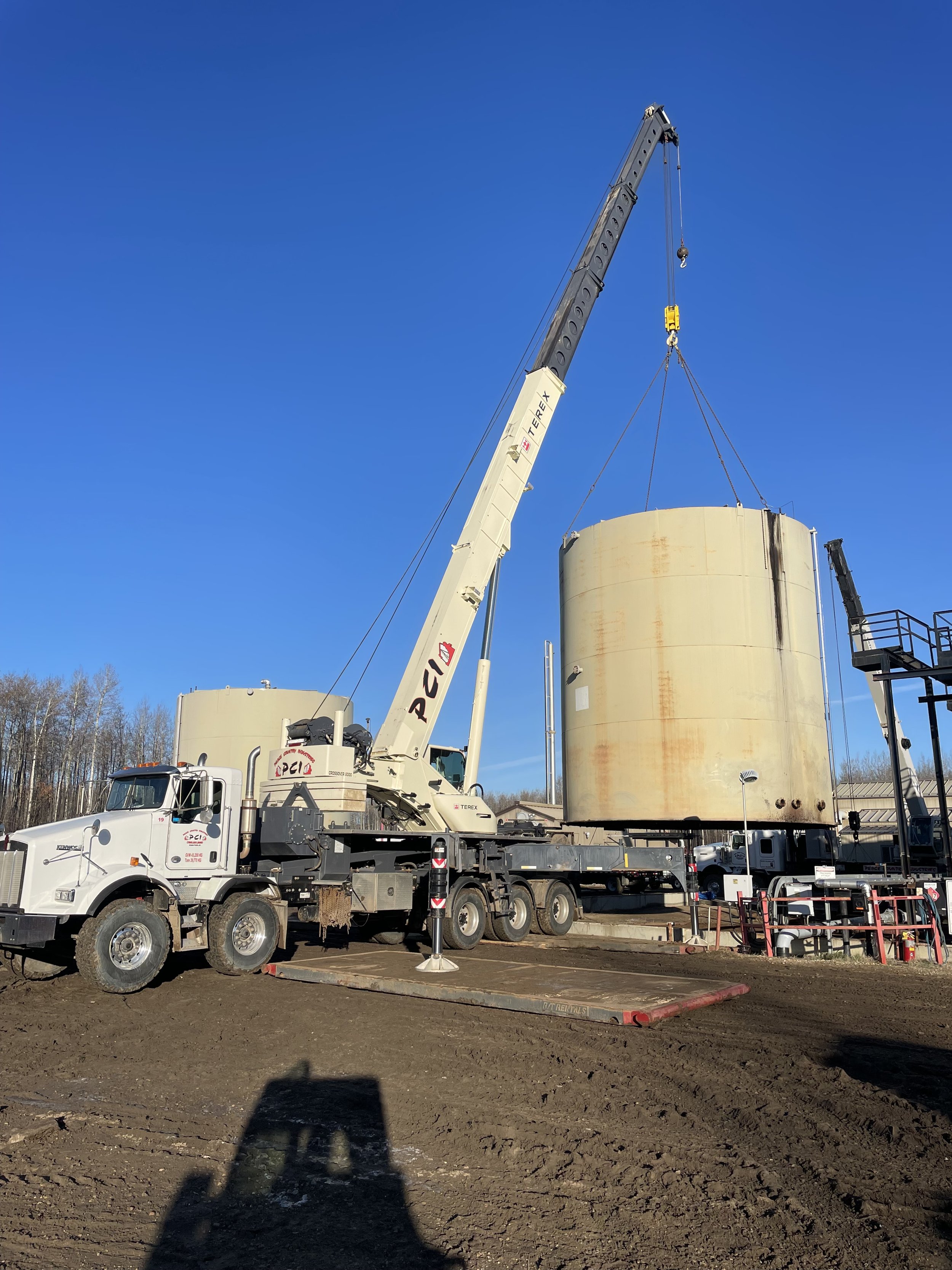 A large crane lifting a beige cylindrical tank at an industrial site on a clear day.
