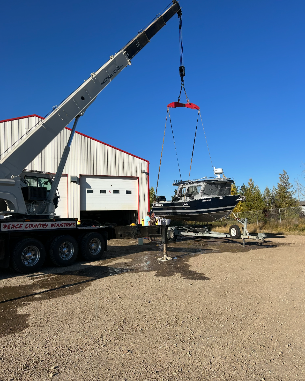 A boat being lifted by a crane onto a trailer in front of a white industrial building with a red trim under a clear blue sky.