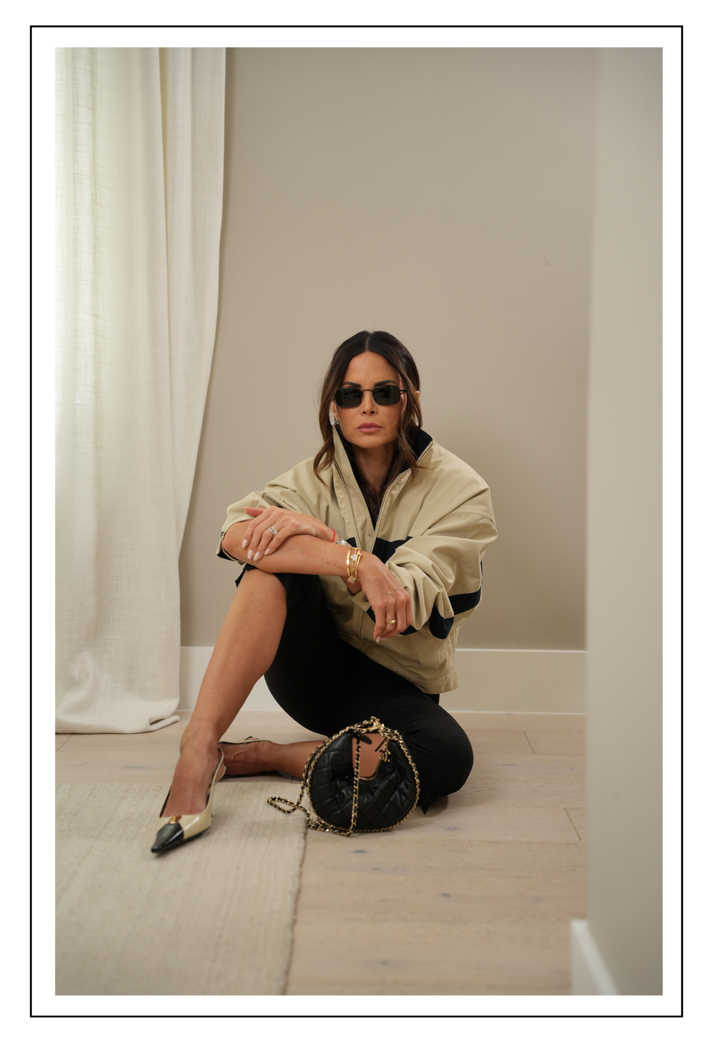 Mujer con gafas grandes, blusa de encaje y falda beige, sosteniendo una bolsa de mano grande con asas negras y decoraciones negras, en interior con pared beige y obra de arte