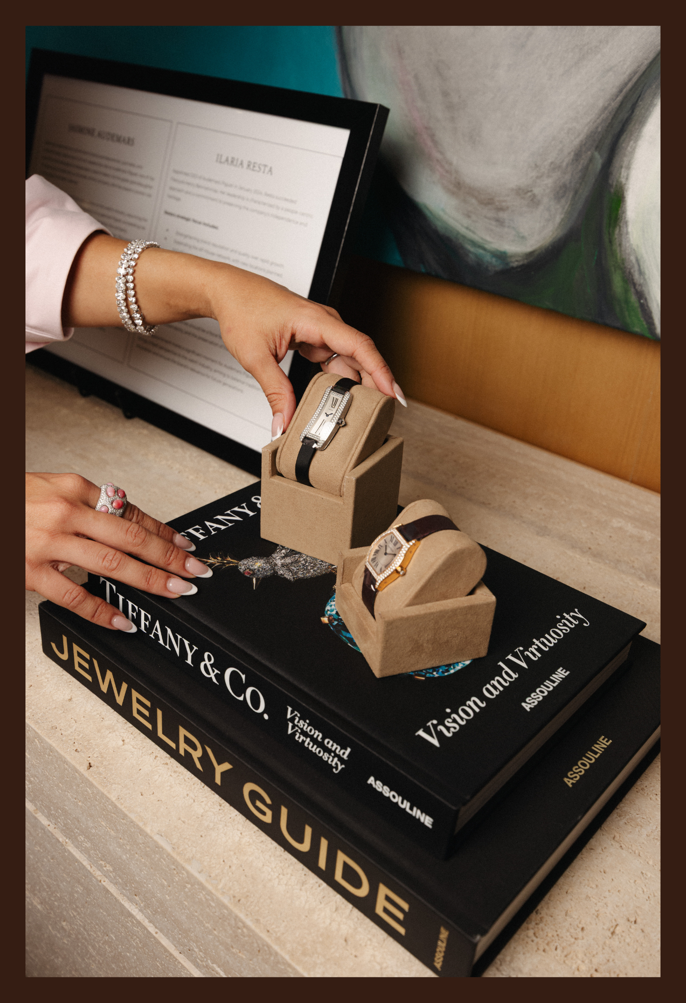 Manos con pulseras y anillos sosteniendo relojes de lujo en una exhibición, con libros de joyería y un cuadro en el fondo.