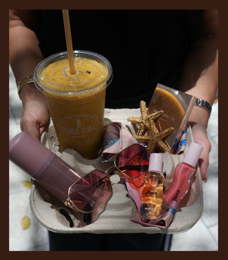 Una persona sostiene una bandeja con un batido amarillo, unas gafas de sol rosadas, dos frascos de perfume, dos estrellas de mar decorativas, y algunos productos de belleza, todo sobre un fondo de arena y superficie arenosa.
