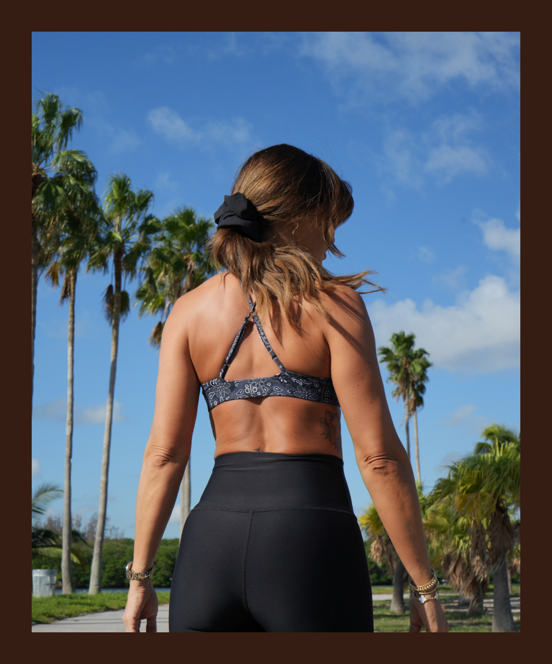 Mujer con cabello al hombro y suelto, usando una blusa deportiva de tirantes y pantalones ajustados negros, de espaldas, rodeada de palmeras y cielo azul con nubes.