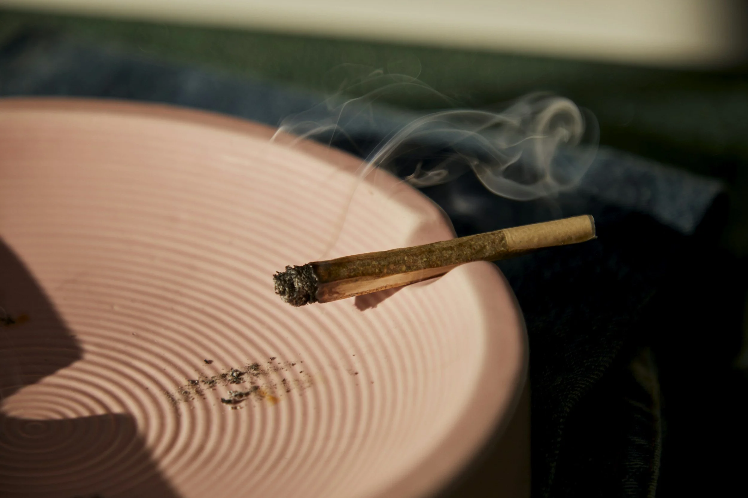 Close-up of a lit incense stick with smoke rising, resting in a pink, textured ashtray with some ash and residual ashes inside.