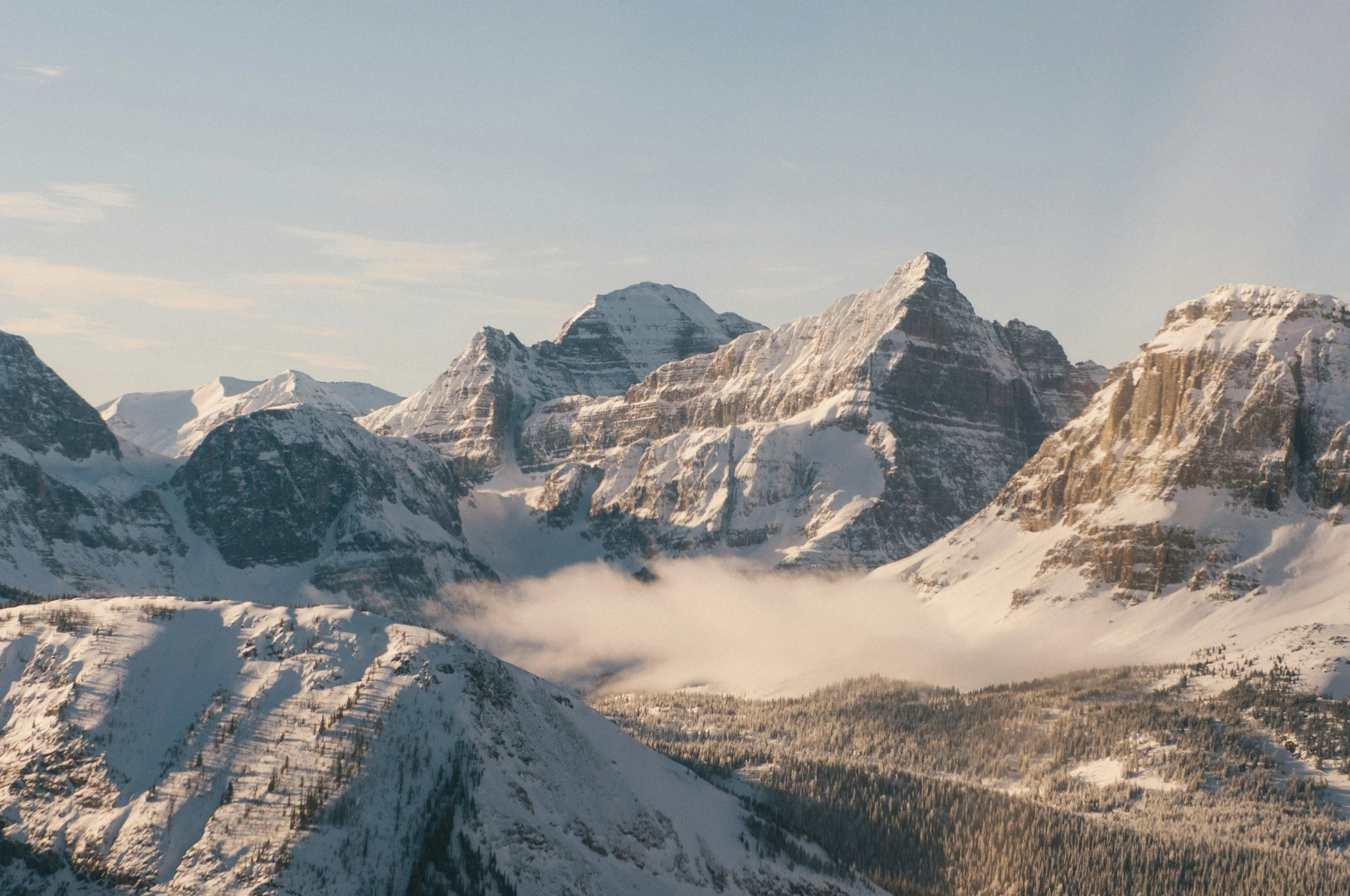 Snow-covered mountain range with steep peaks, clouds in a valley, and a forested hillside in the foreground.
