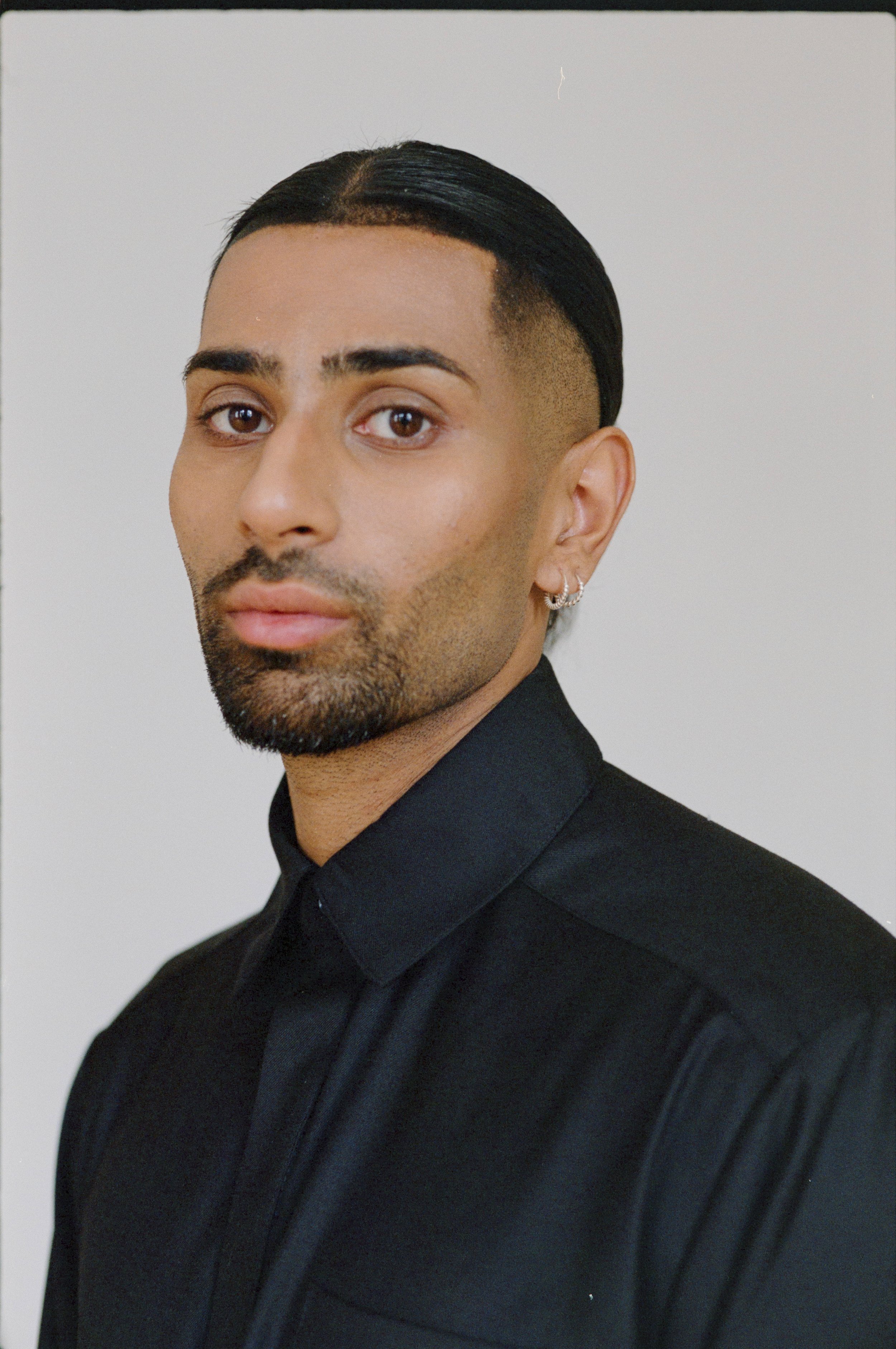 A person with a neatly trimmed beard and slicked-back hair, wearing earrings and a black button-up shirt, looking directly at the camera against a plain background.