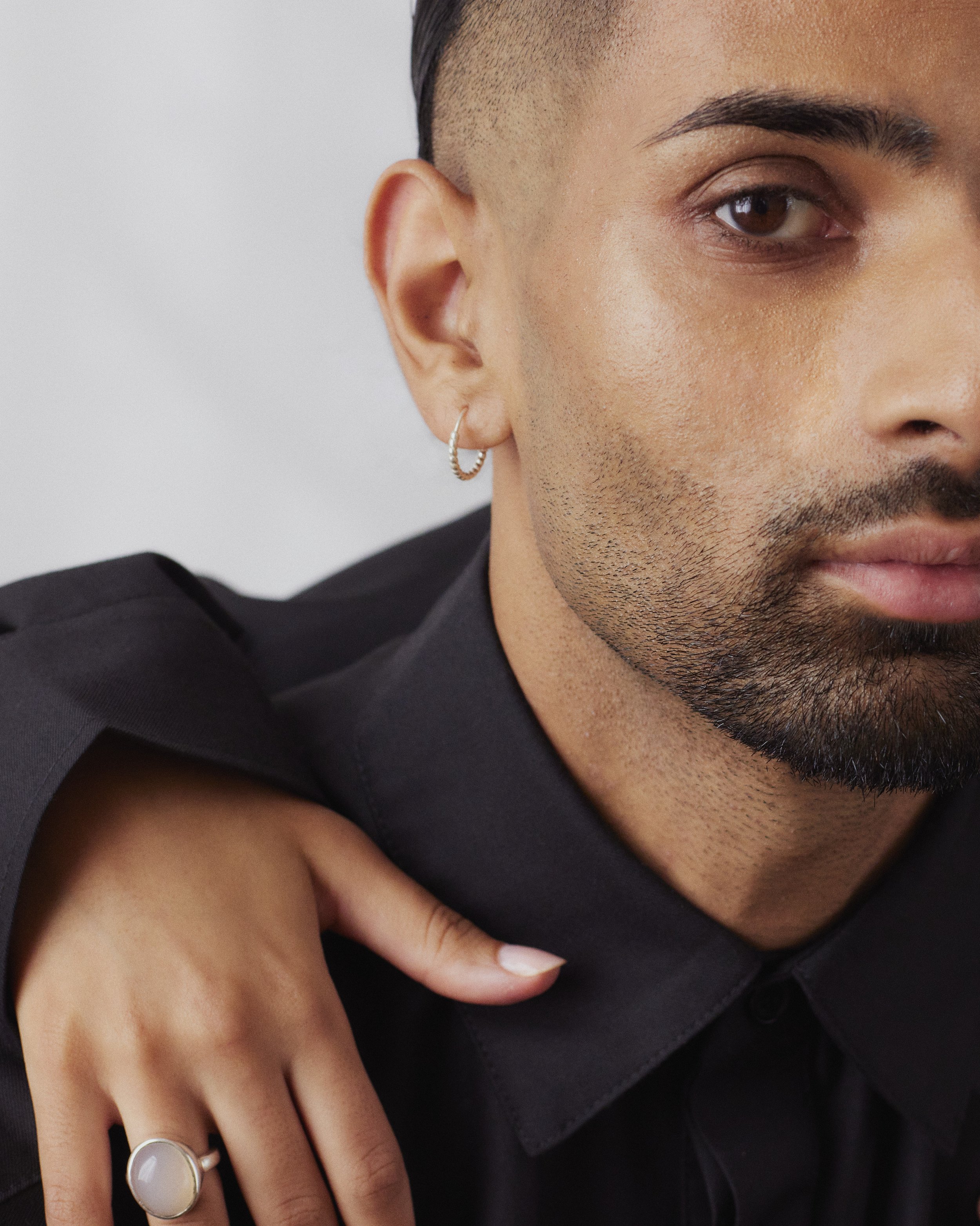 Close-up of a man wearing a black shirt and hoop earring, with a hand resting on his shoulder featuring a ring with a round stone.