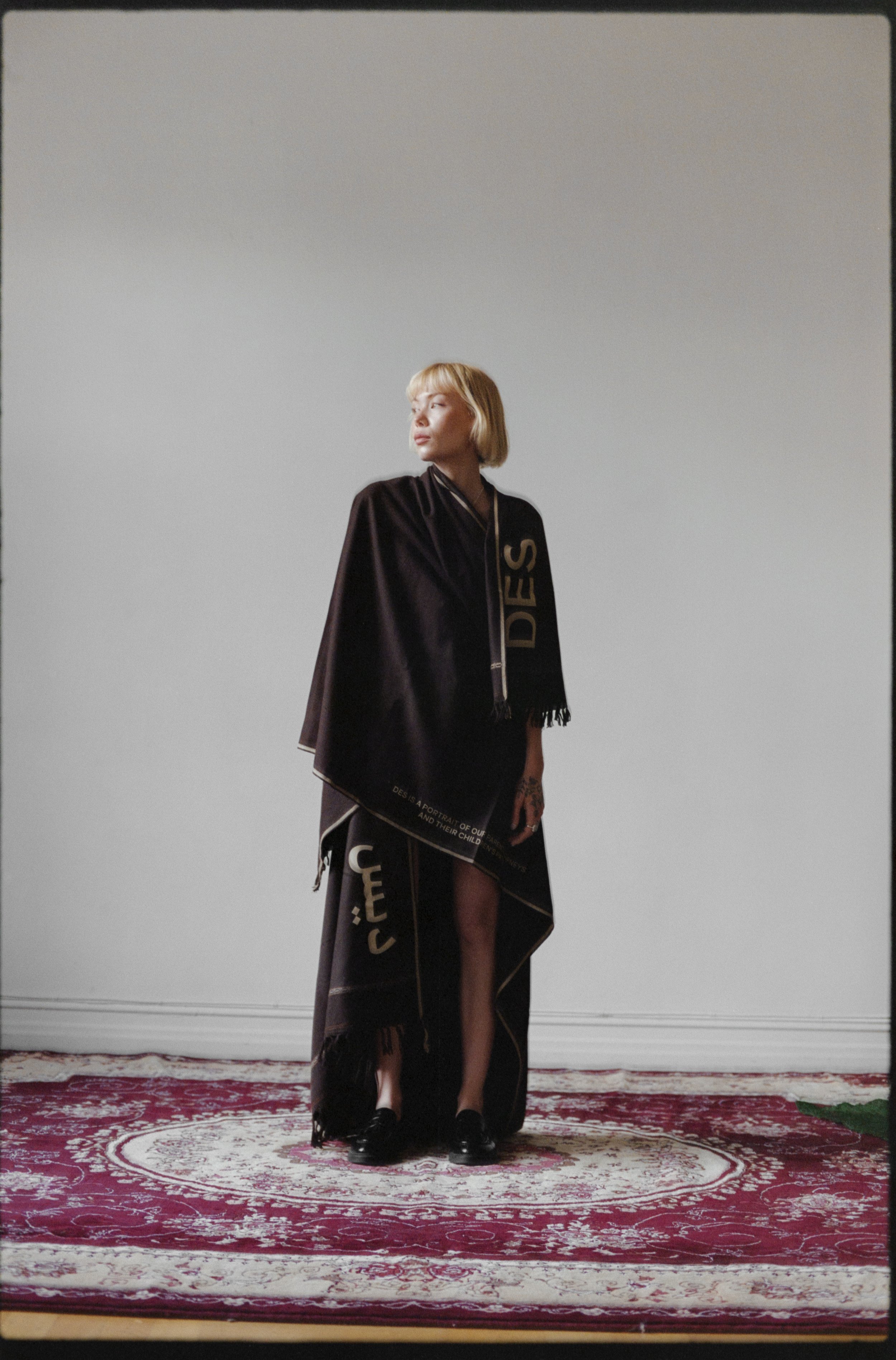 Person wearing a dark brown poncho with text, standing on a patterned rug in a white-walled room.