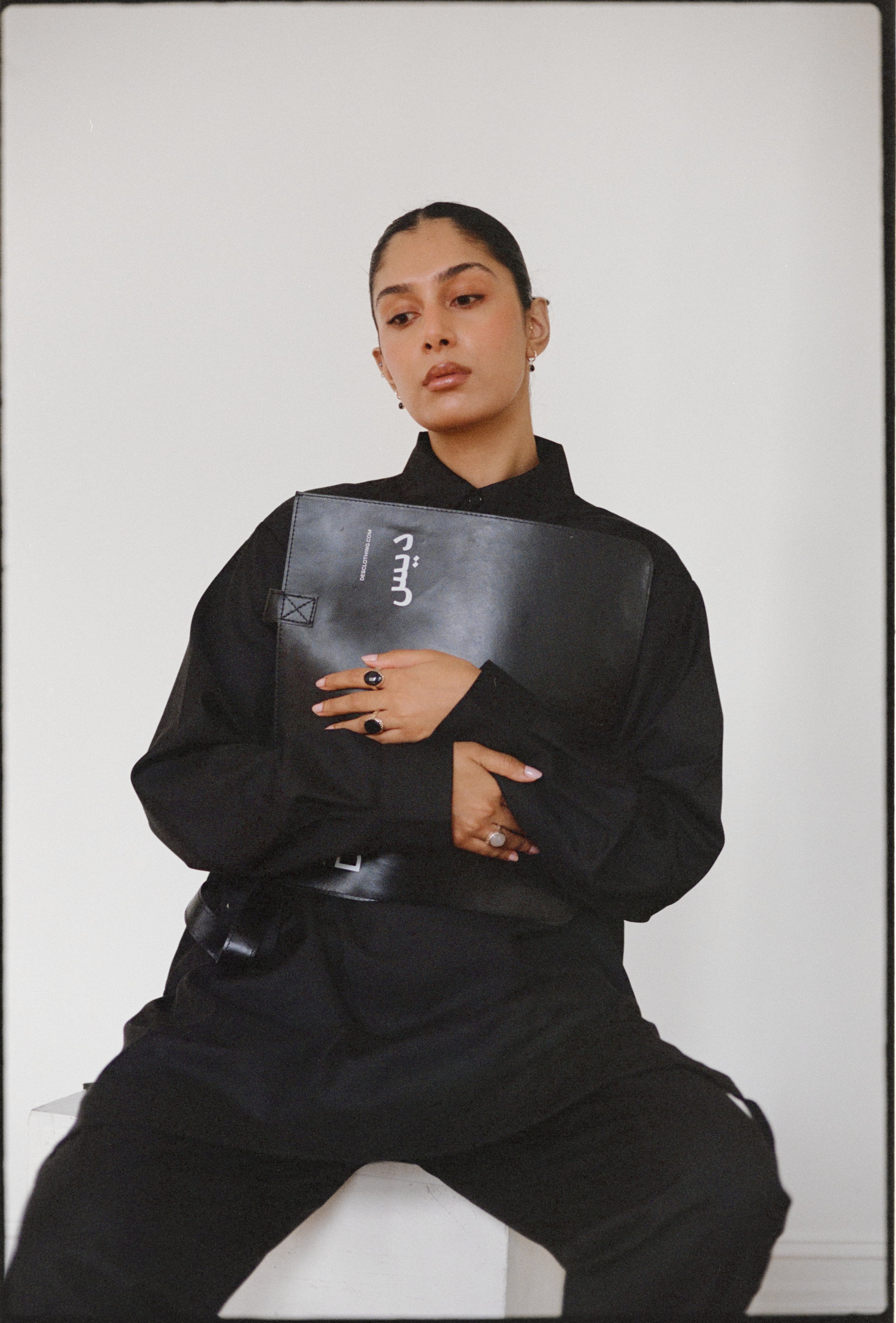 Person in black outfit holding a black portfolio, sitting against a plain white background.