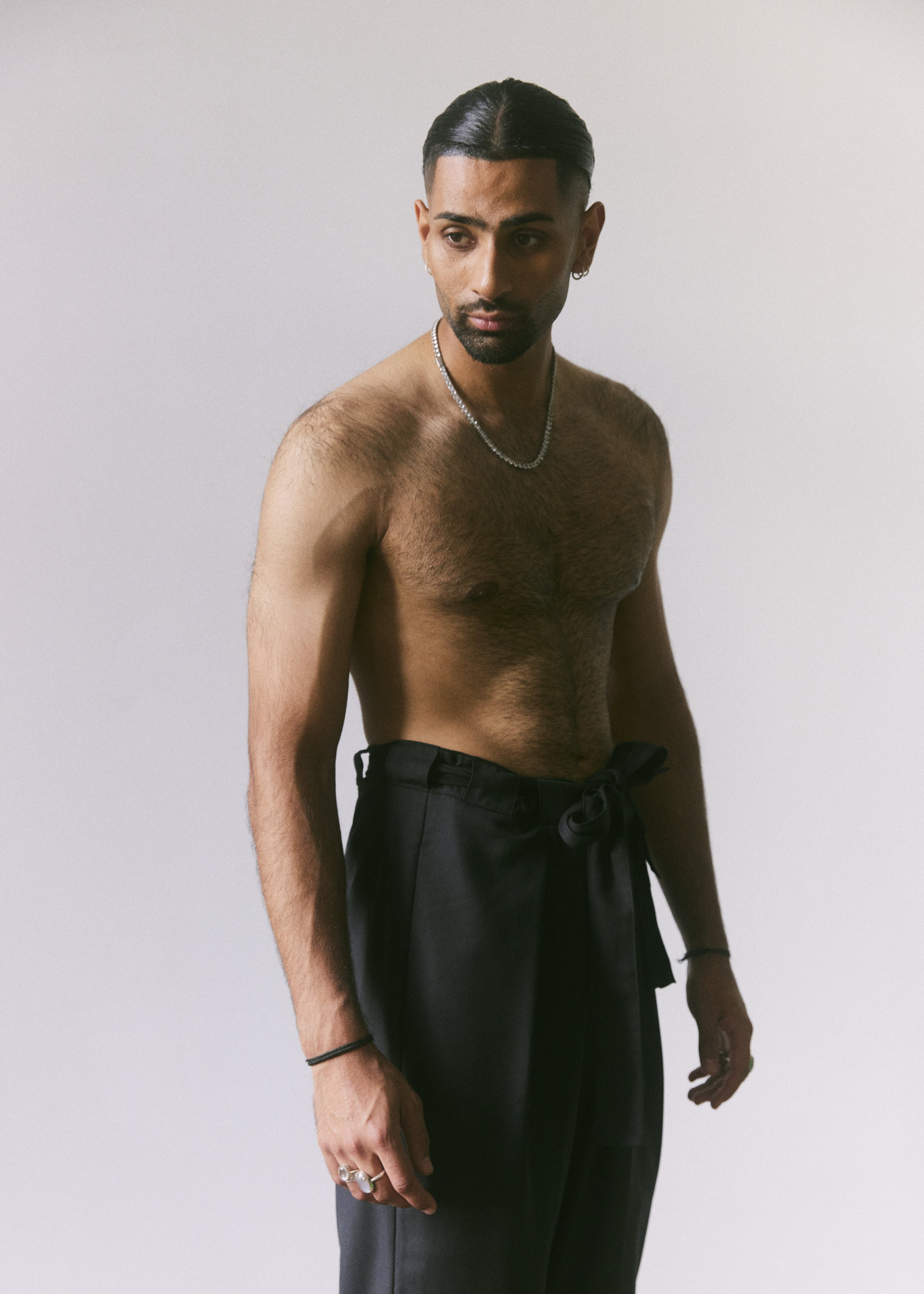 Shirtless man with slicked-back hair wearing black pants and jewelry, standing against a plain background.