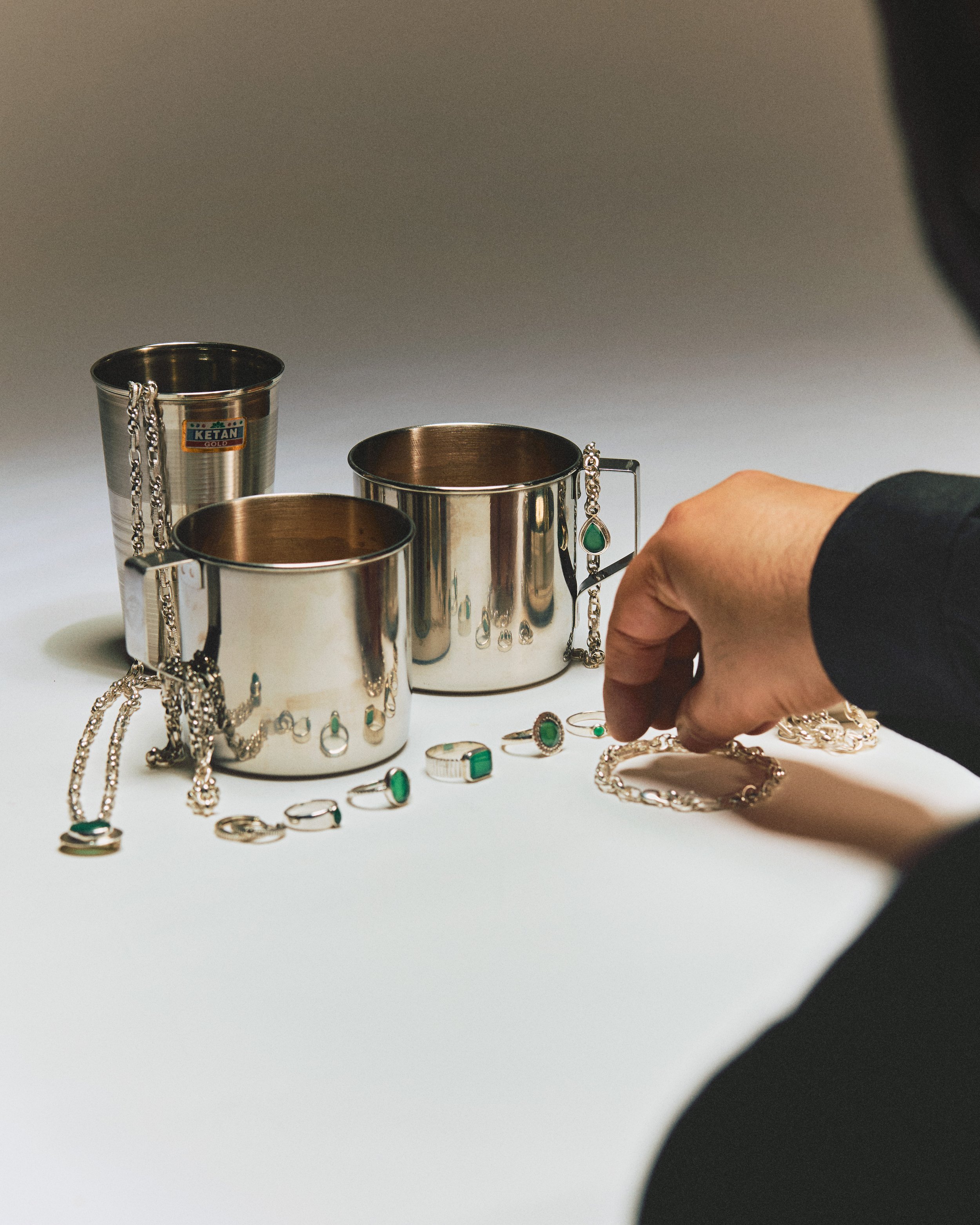 Silver cups and jewelry on a white surface with a hand arranging rings and necklaces.