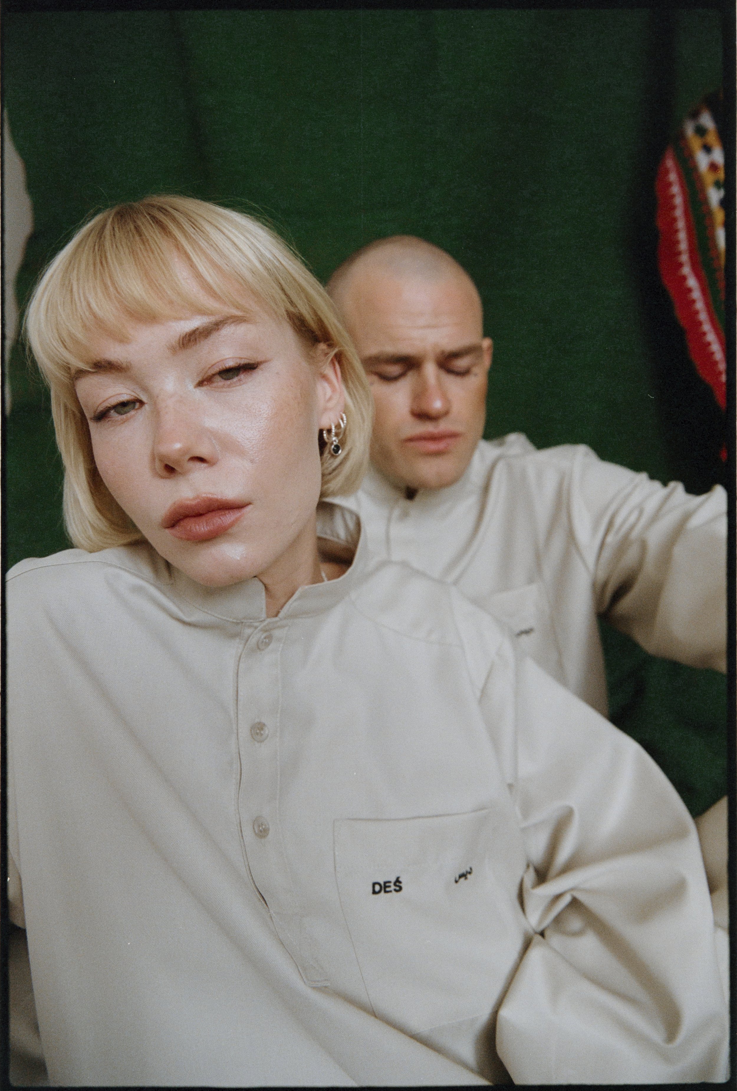 Two people wearing matching white shirts pose in front of a green backdrop. The person in the foreground has short blond hair and appears close to the camera, while the person behind has a shaved head. They both have neutral expressions.