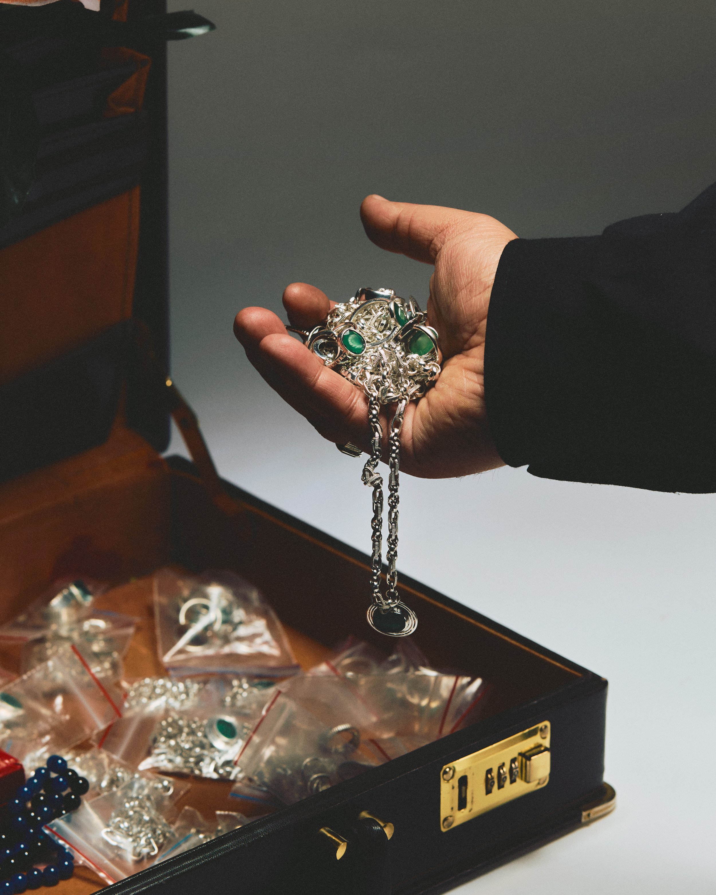 Hand holding a silver bracelet with green stones over an open box filled with bagged jewelry items.
