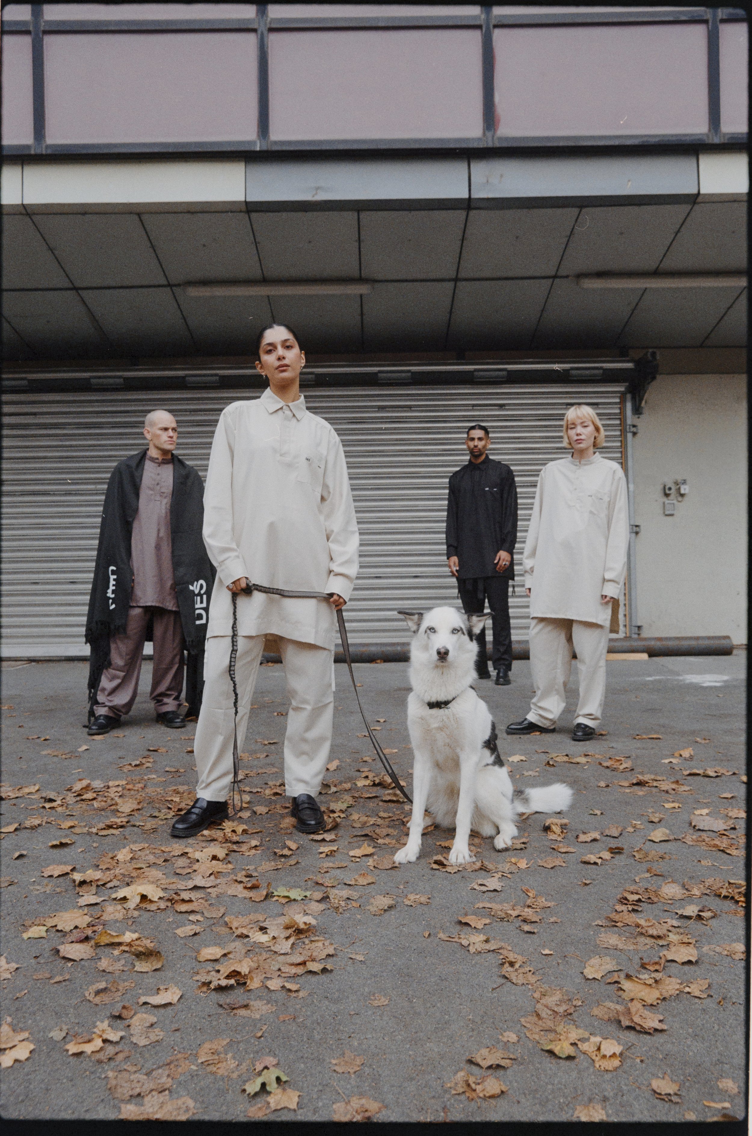 Group of people in fashion outfits with a white and black dog in an urban setting with fallen leaves.