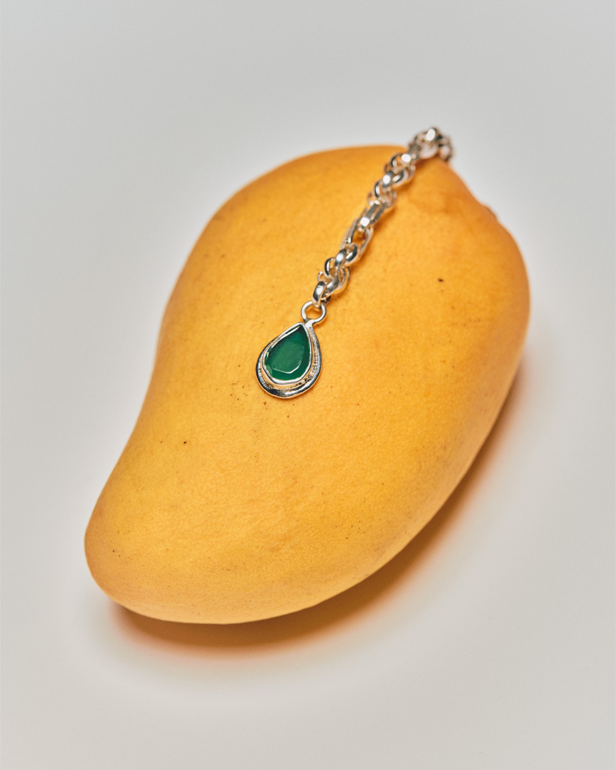 A ripe mango with a silver chain bracelet featuring a green teardrop pendant resting on it.