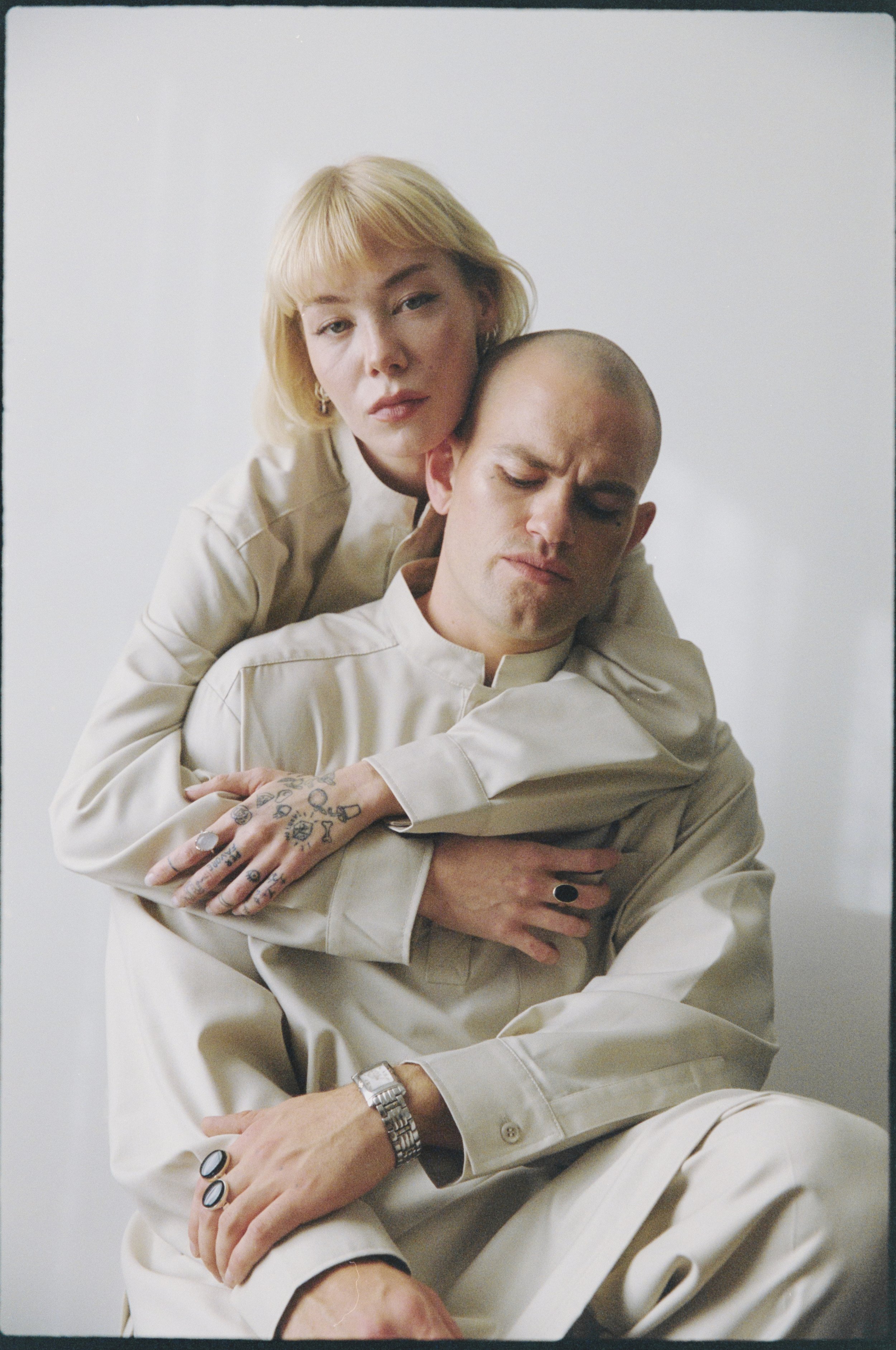 A woman with short blond hair and a man with a shaved head, both in light-colored clothing, embracing each other. The woman has tattoos on her hand and is wearing rings. The man is wearing a watch and multiple rings.