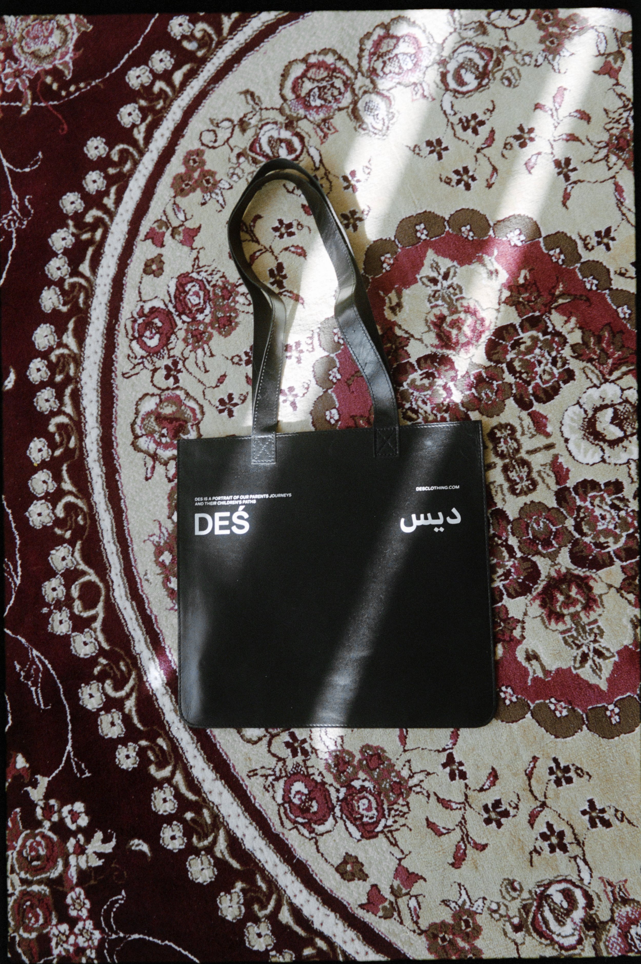 Black tote bag with 'DES' text on a floral patterned rug.