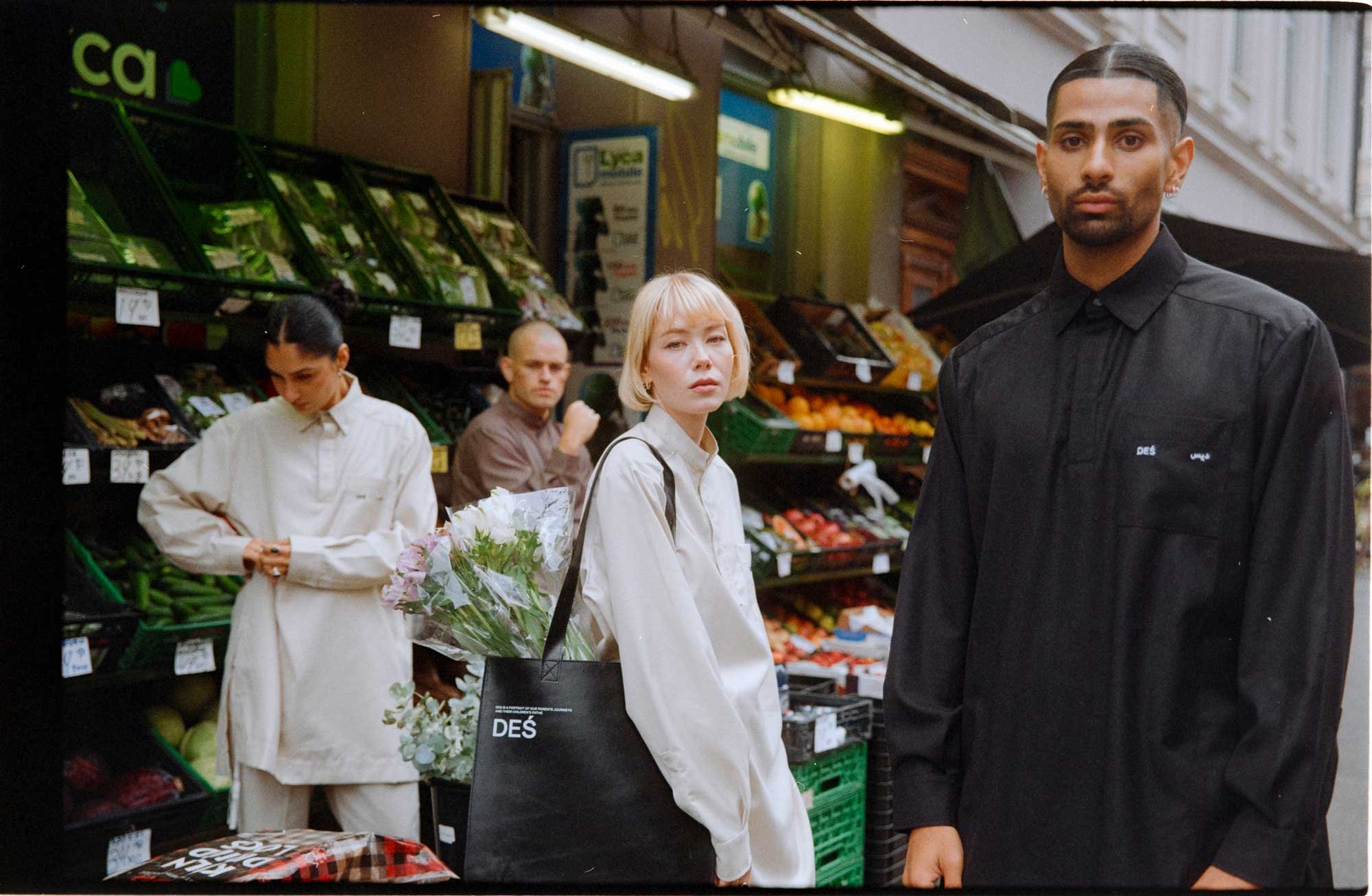 People in casual clothing shopping at an outdoor market with fruits and vegetables in the background. One person carries a black tote bag with flowers.
