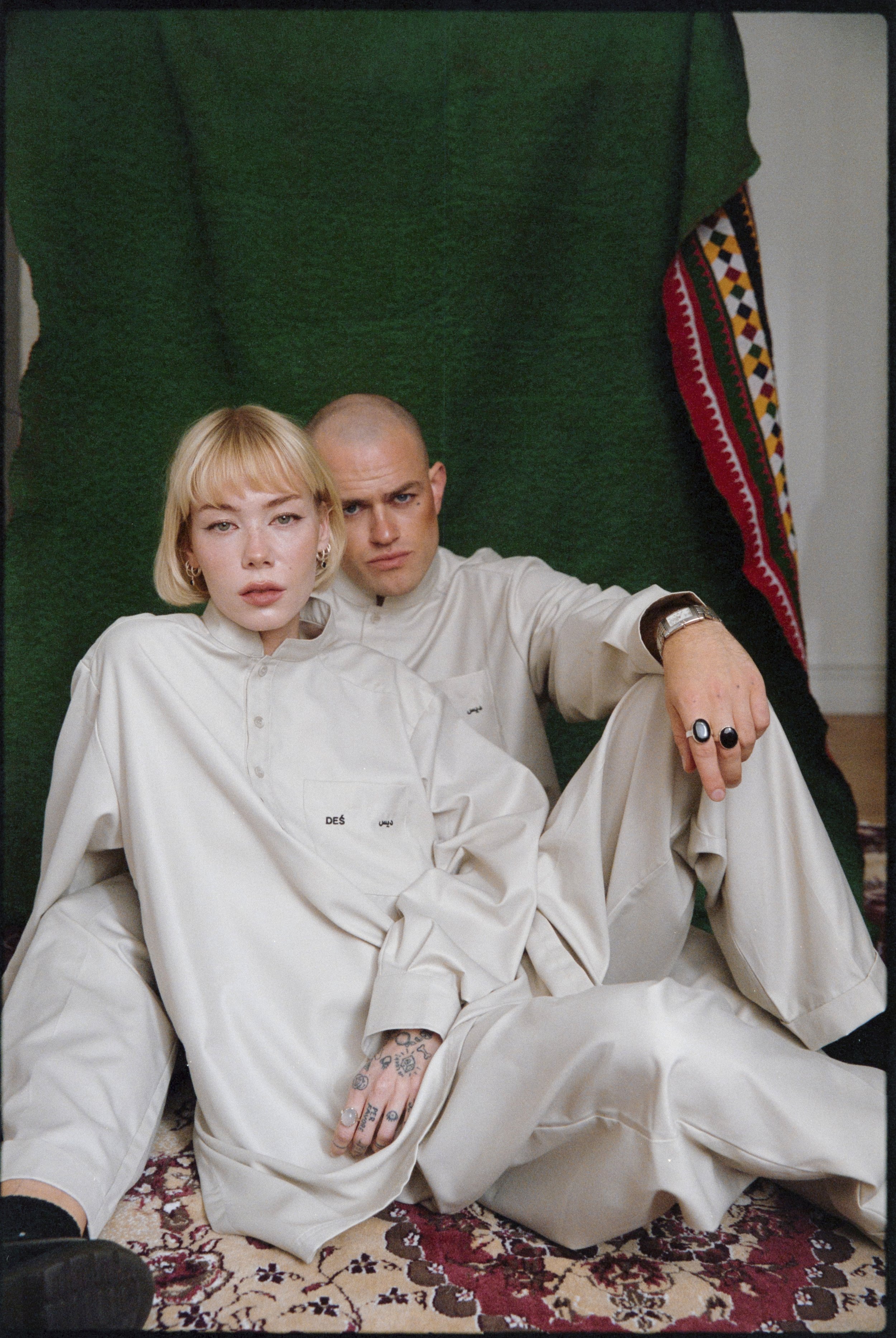 Two people sitting on a patterned rug, wearing matching light beige outfits, with a green backdrop and a colorful textile on the side.