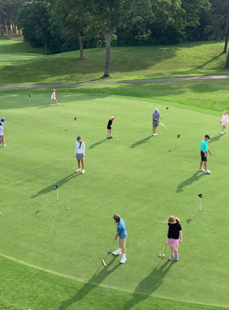People practicing putting on a golf green, surrounded by trees and grass.