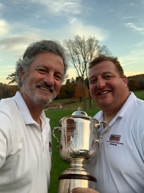 Two men in white shirts smiling and holding a large silver trophy outdoors on a grassy area with trees in the background.