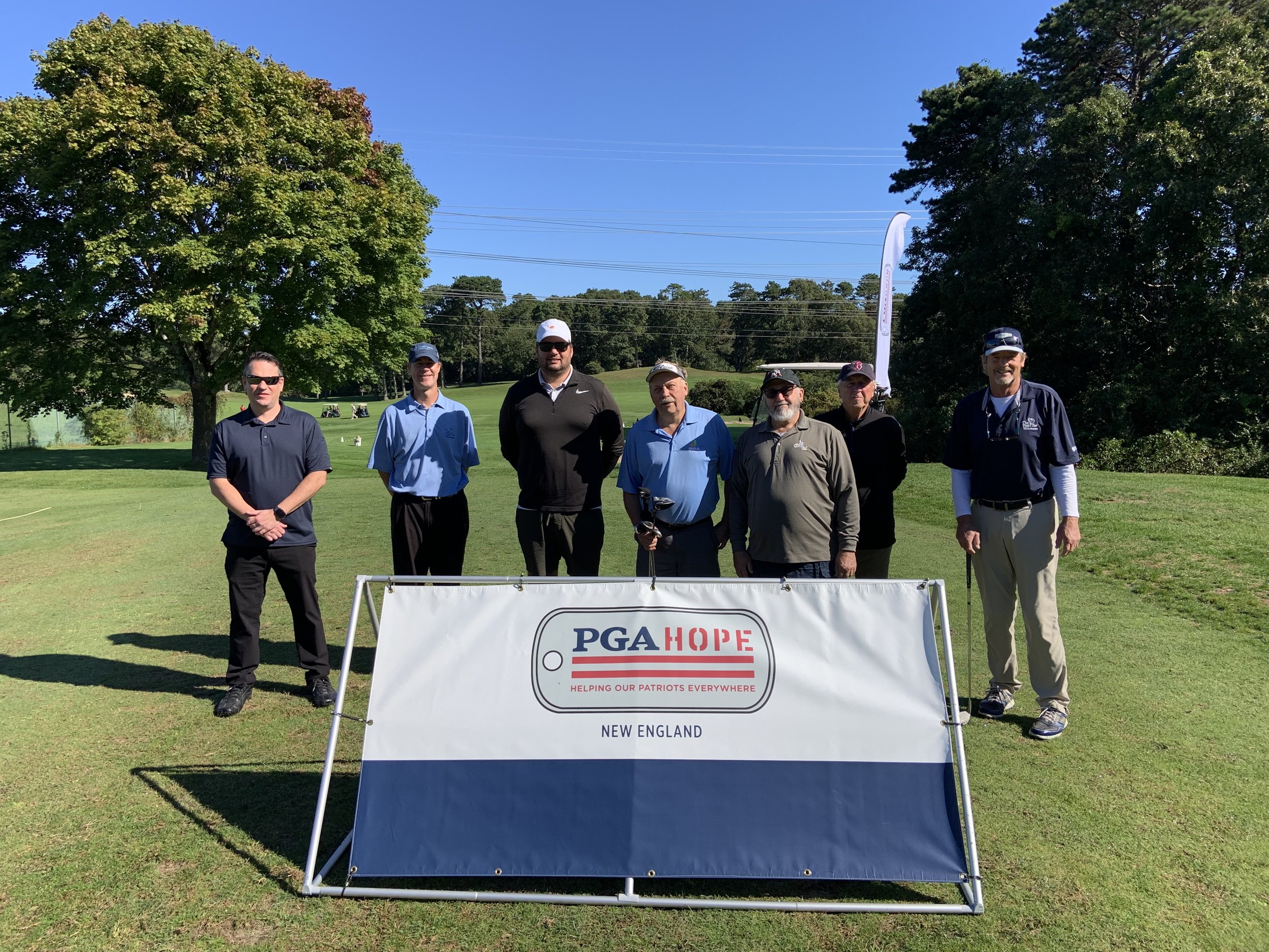 Group of seven men standing on a golf course behind a PGA HOPE New England sign.