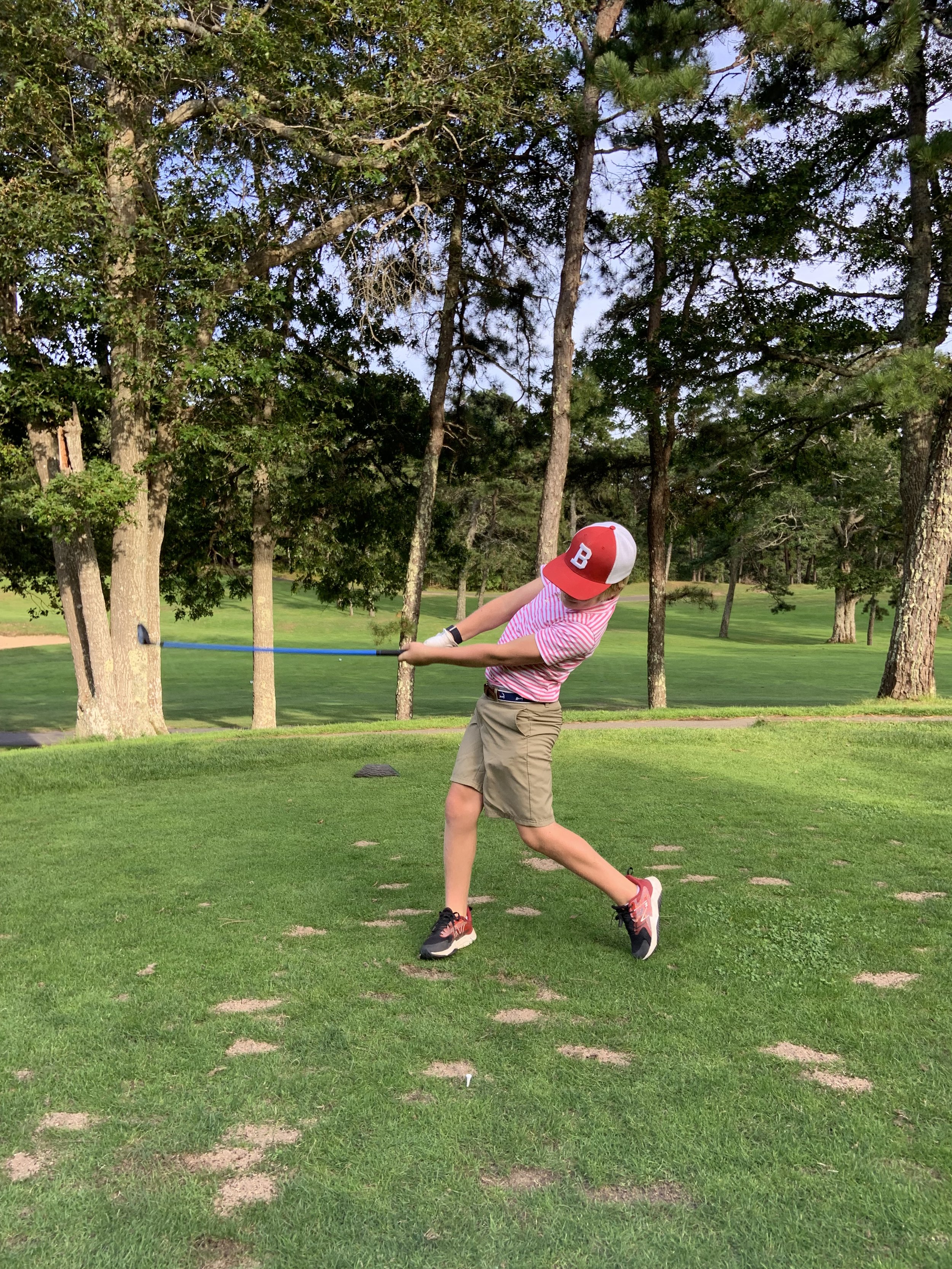 Child swinging a golf club on a grassy course with trees in the background.