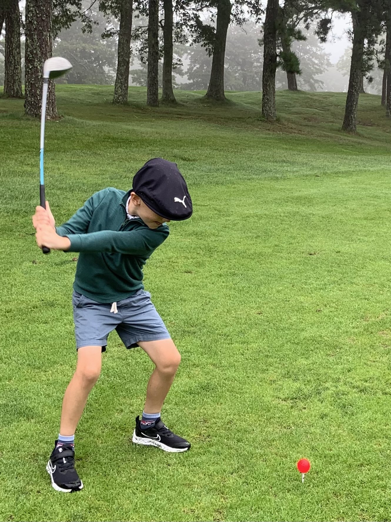 Young golfer swinging club on a golf course, ball on tee, surrounded by trees.