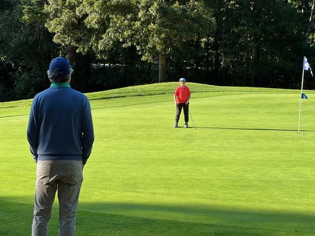Two people on a golf course, one wearing a red jacket near the hole, the other in a blue jacket standing further back.