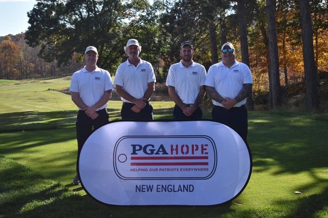 Four men in white shirts and hats stand outdoors on a golf course next to a sign saying "PGA Hope Helping Our Patriots Everywhere New England." The background includes trees and grass, suggesting a sunny day.