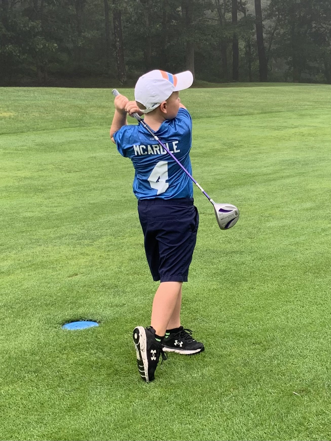 A young child wearing a blue jersey and white cap swings a golf club on a lush green golf course.