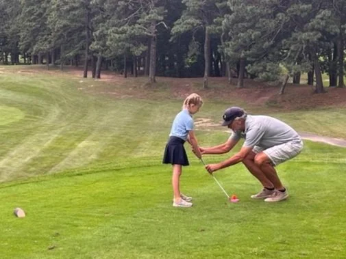 A young girl learning golf with instructor Coach Dave on a green course. The instructor is crouched down helping the girl with her grip on the club. Trees are visible in the background.