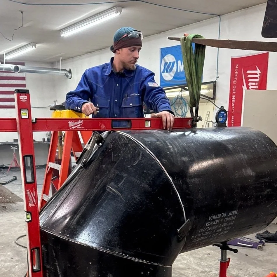 Man wearing a blue work shirt and a beanie, standing on a ladder, measuring a large black pipe in a workshop.