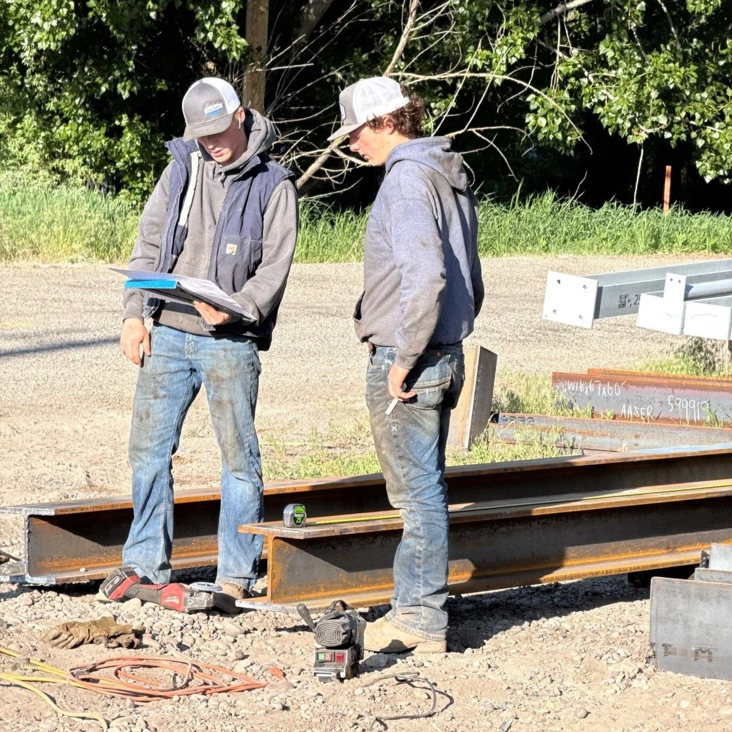 Two construction workers standing on a steel beam at a construction site, reviewing blueprints, with tools and materials around them.