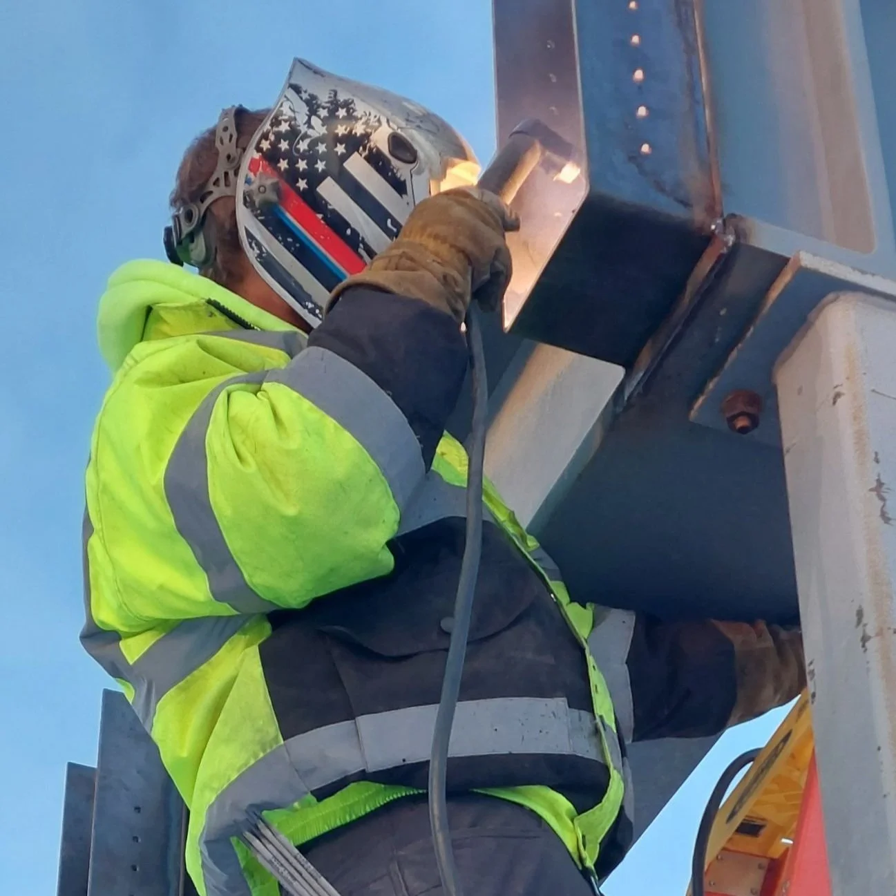 A construction worker wearing a protective helmet with an American flag design, a high-visibility yellow jacket with gray accents, and gloves, is welding or working on a steel structure outdoors during dusk or dawn.