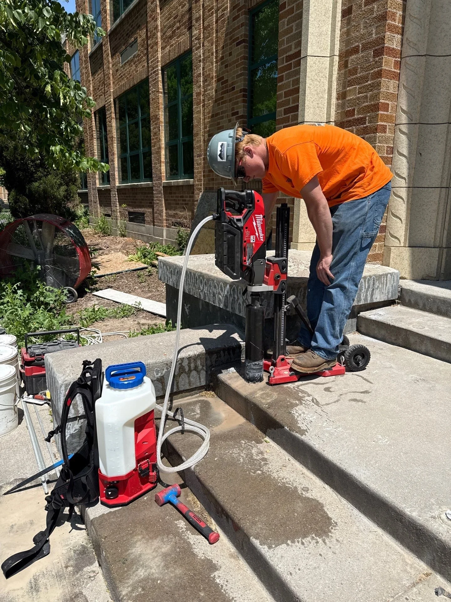 A worker using a core drill to cut into concrete steps outside a brick building on a sunny day.