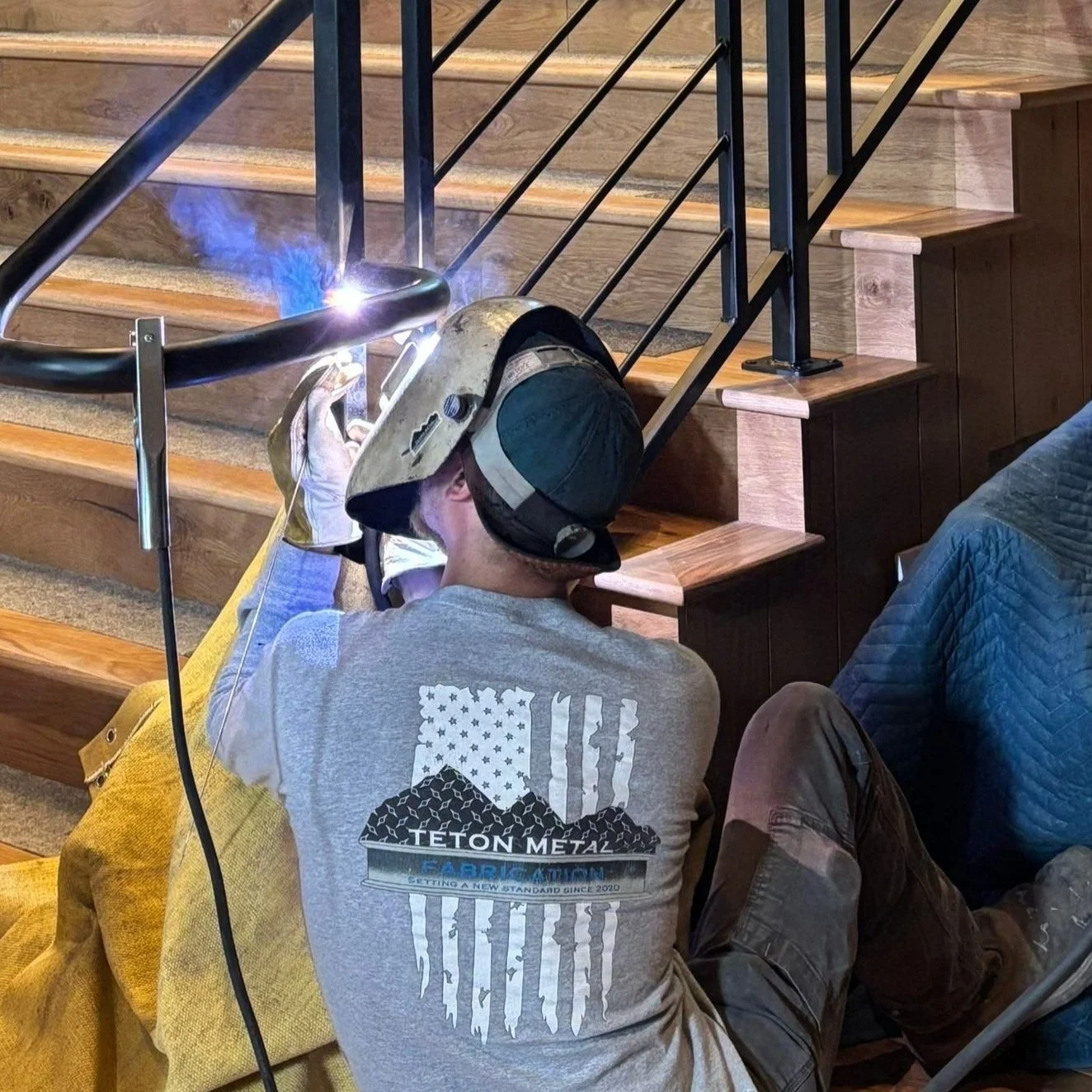 A person welding on a staircase railing, wearing a welding helmet and work gloves, with protective gear, inside a building with wooden stairs and black metal railing.