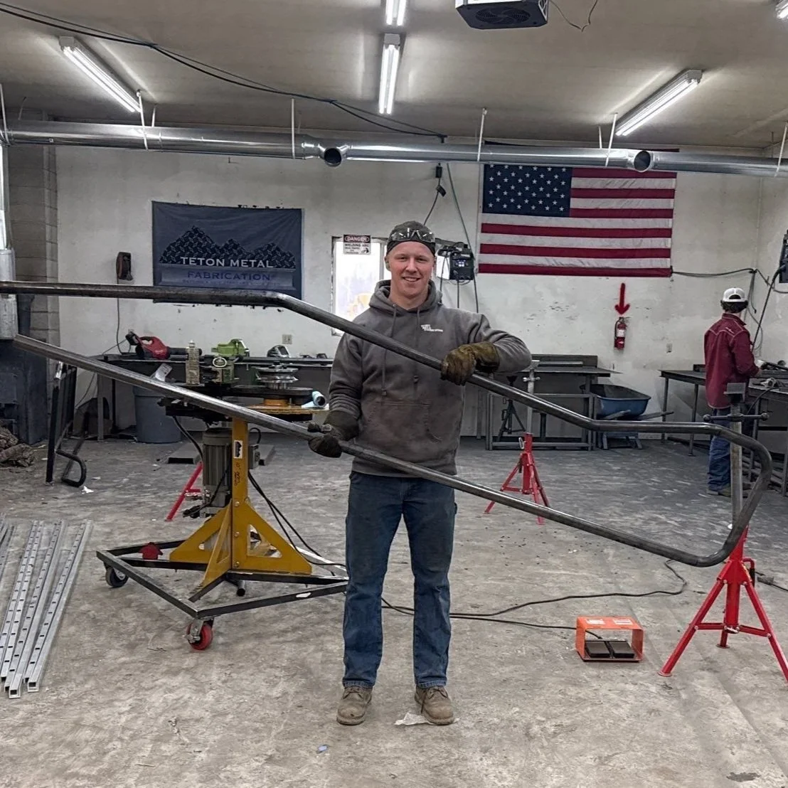 A man in a workshop holds a large metal frame while smiling at the camera. There are tools, workbenches, an American flag, and a Teton Metal Fabrication banner in the background.