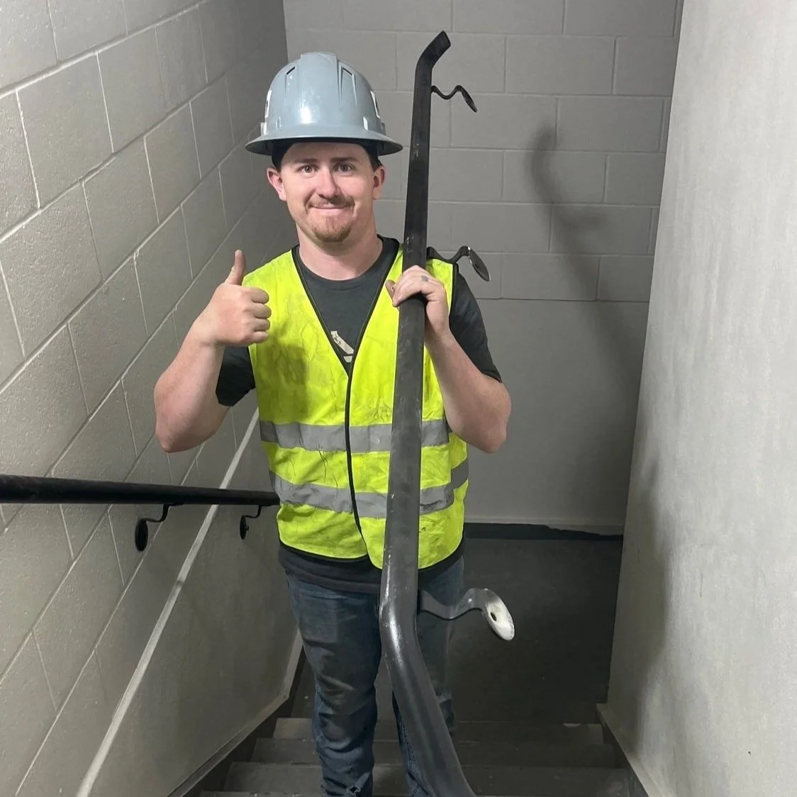 A construction worker wearing a gray helmet, yellow safety vest, and black t-shirt, smiling and giving a thumbs-up while holding a crowbar, standing on a stairwell with beige brick walls.
