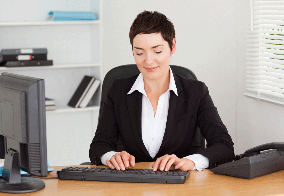 A woman in a business suit working at a desk with a computer, telephone, and shelves in the background.