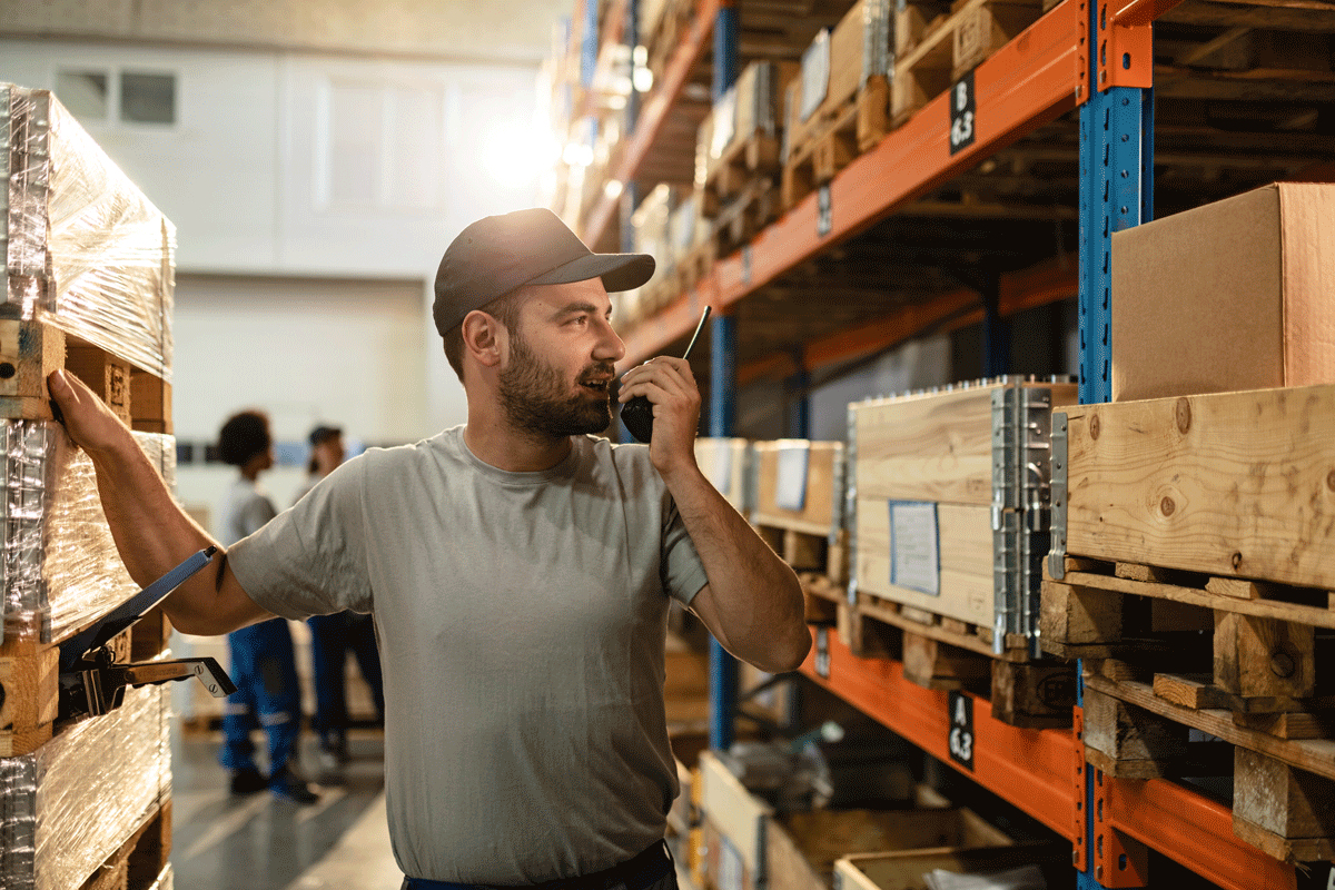A man in warehouse holding a walkie-talkie, looking at wooden pallets and boxes on metal shelves.