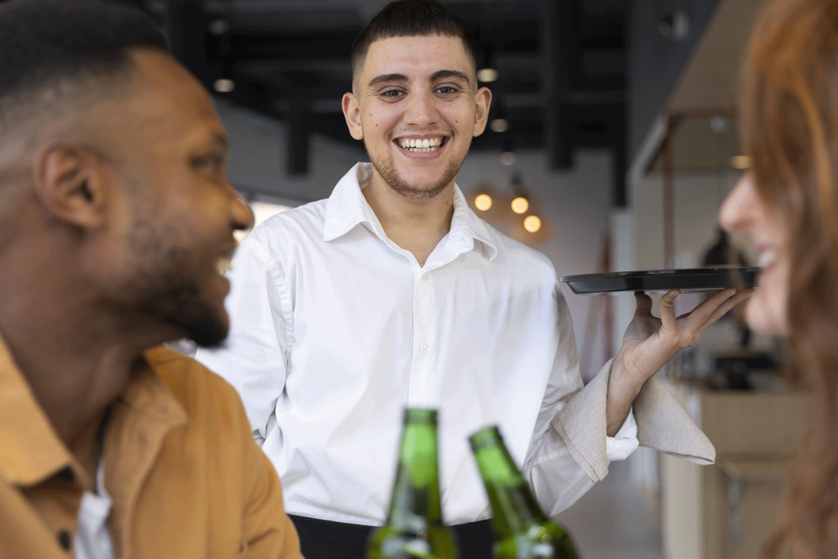 A smiling waiter in a white shirt serving drinks to a couple at a restaurant.