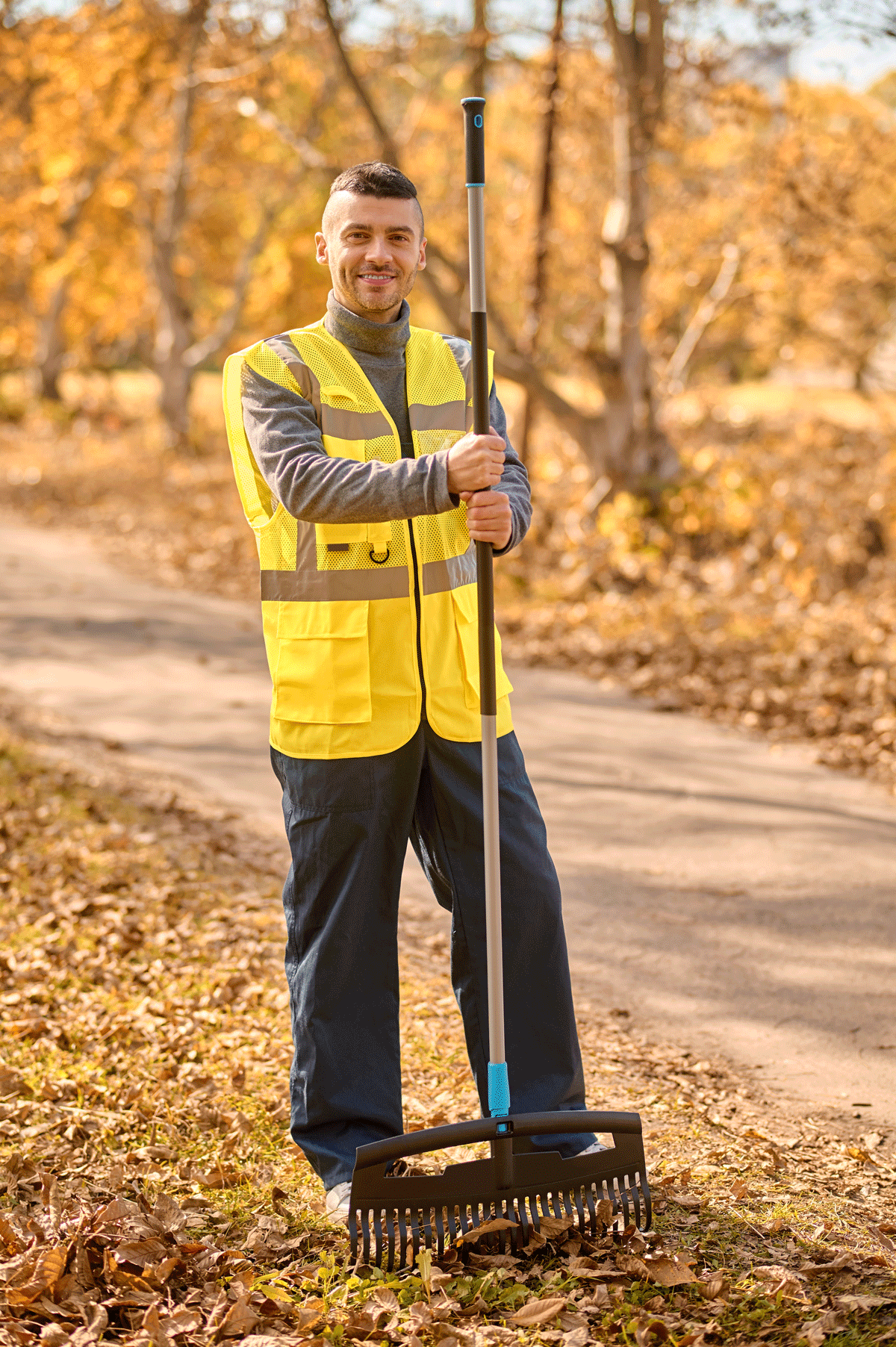 A man in a yellow safety vest and dark pants holding a leaf rake, standing on a leaf-covered dirt path in a park with autumn foliage.