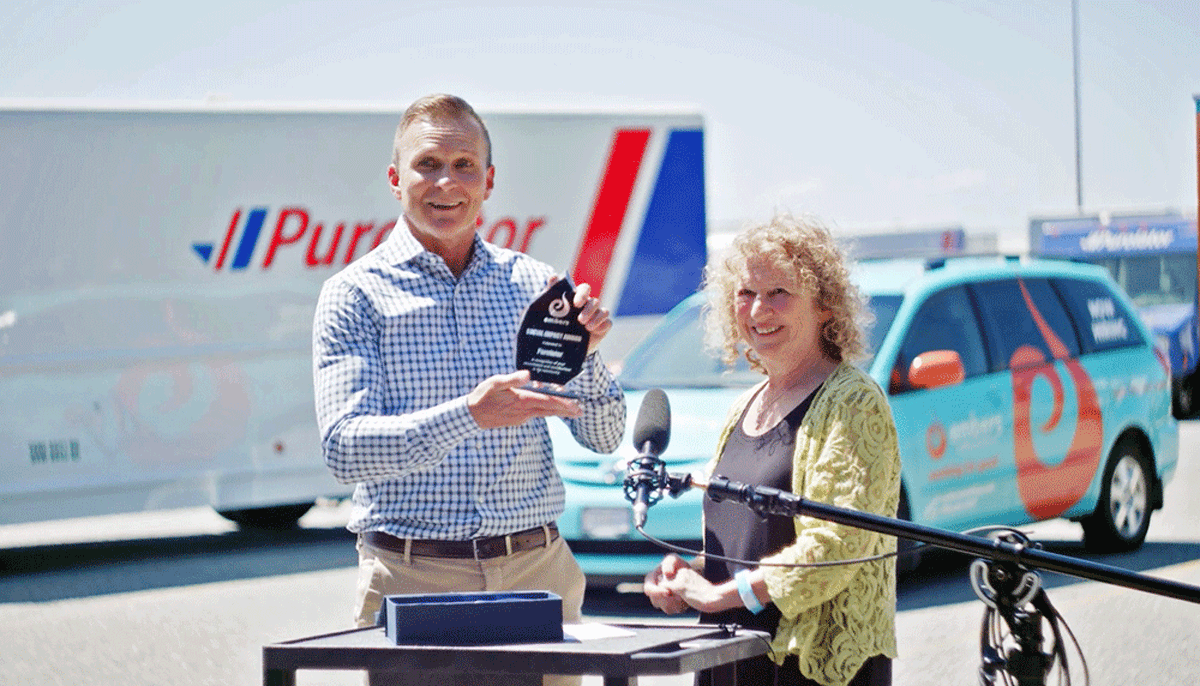 A man and woman standing outdoors near vehicles, with the man holding a trophy or award, smiling at the camera.