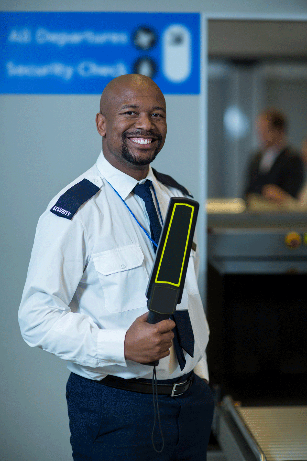 Smiling security officer in uniform at airport security checkpoint holding a handheld scanner.