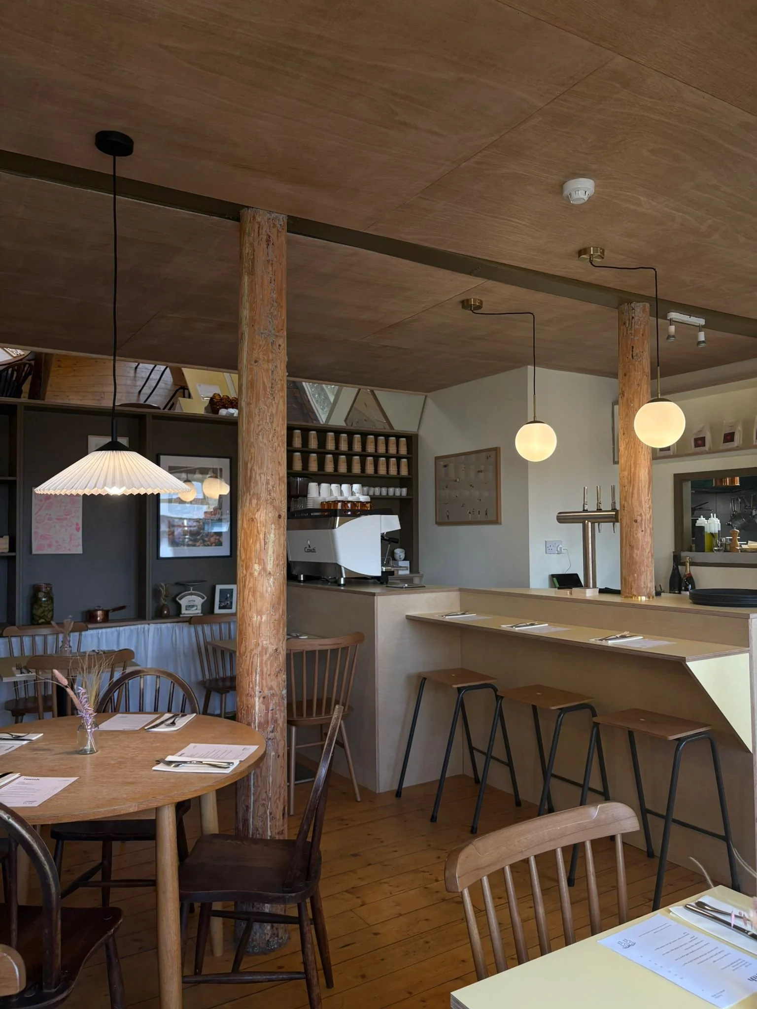Interior of a cozy restaurant with wooden tables and chairs, pendant lights, and a bar area with stools and shelves of cups and plates.