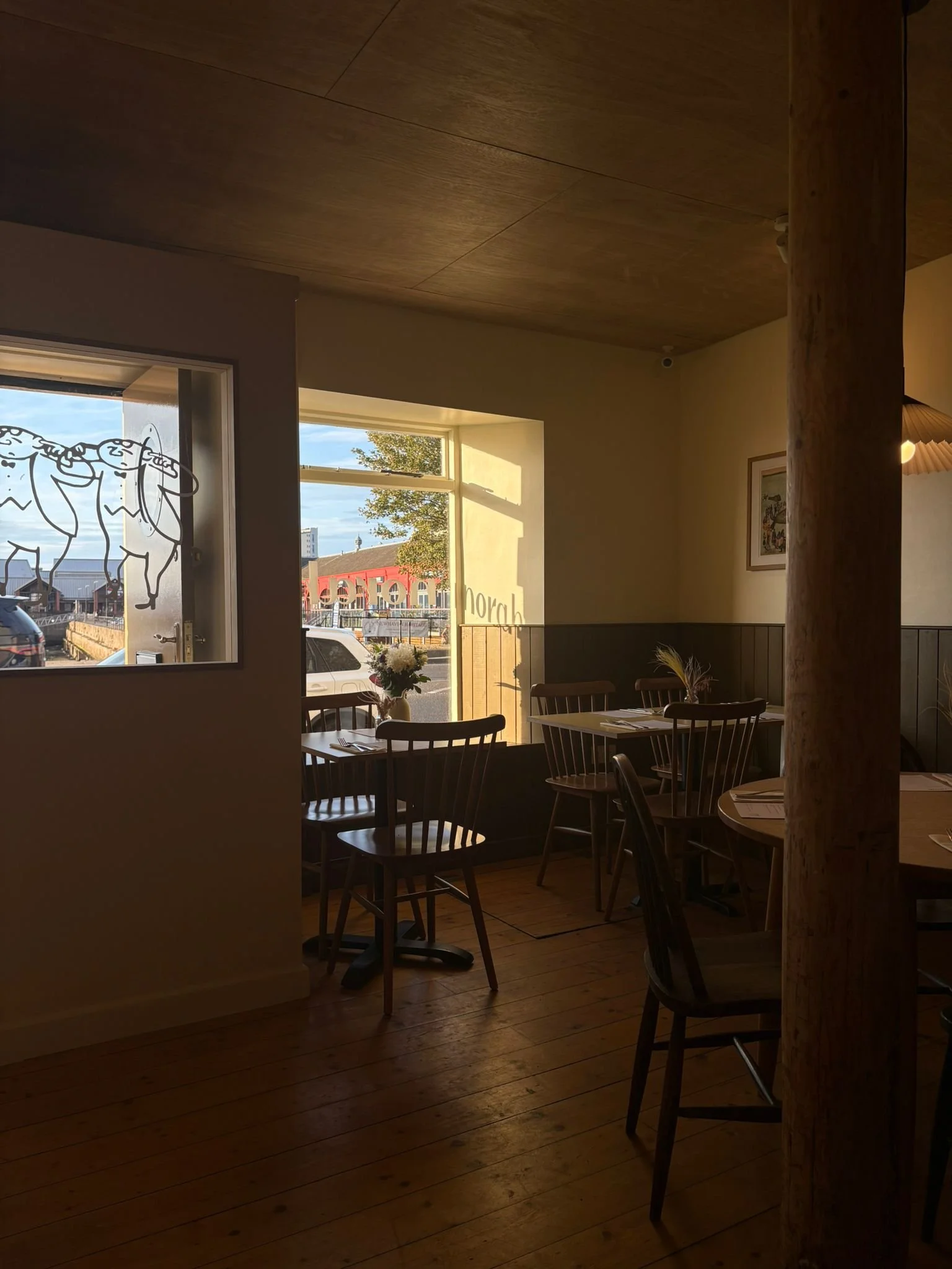 Interior of a cozy restaurant with wooden floors and furniture, sunlight streaming through large windows, and a small vase of flowers on a table near the window.