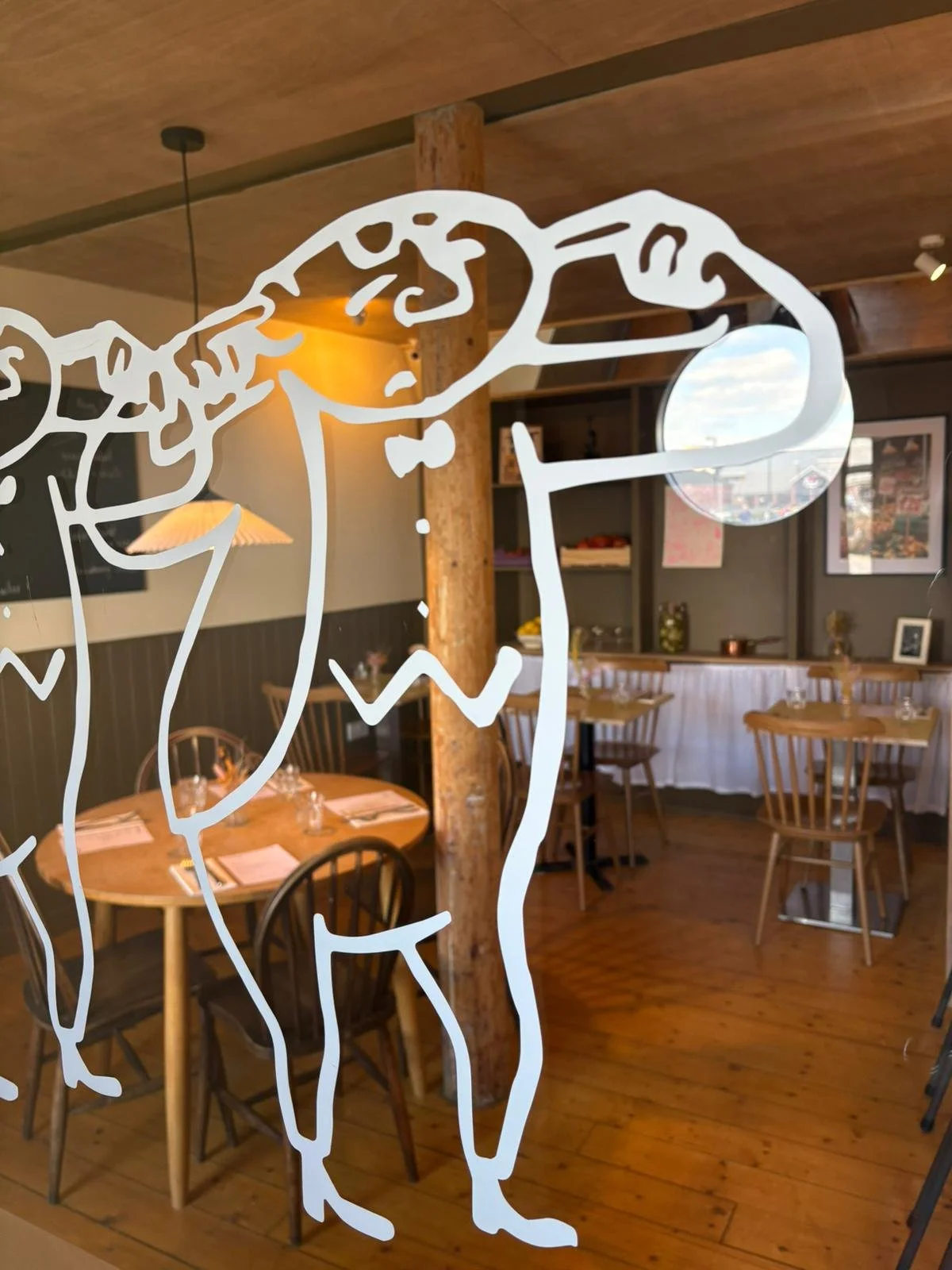 Interior of a cozy restaurant with wooden tables and chairs, decorated with artwork on the walls, seen through a glass with a white line drawing of a dog.