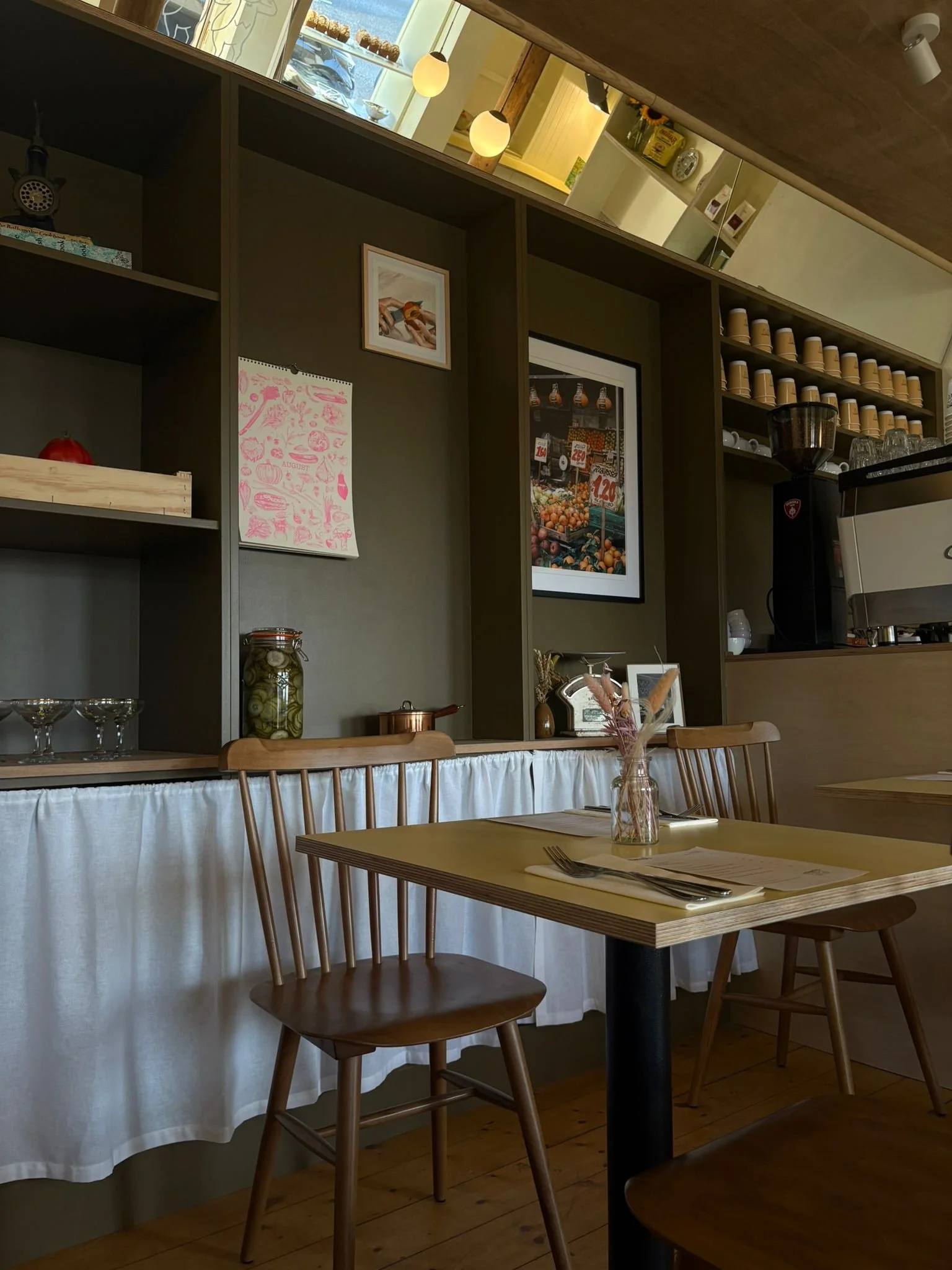 Interior of a cozy cafe with a wooden table, two wooden chairs, a bookshelf with framed pictures, jars, and decorative items, and a counter with coffee equipment.