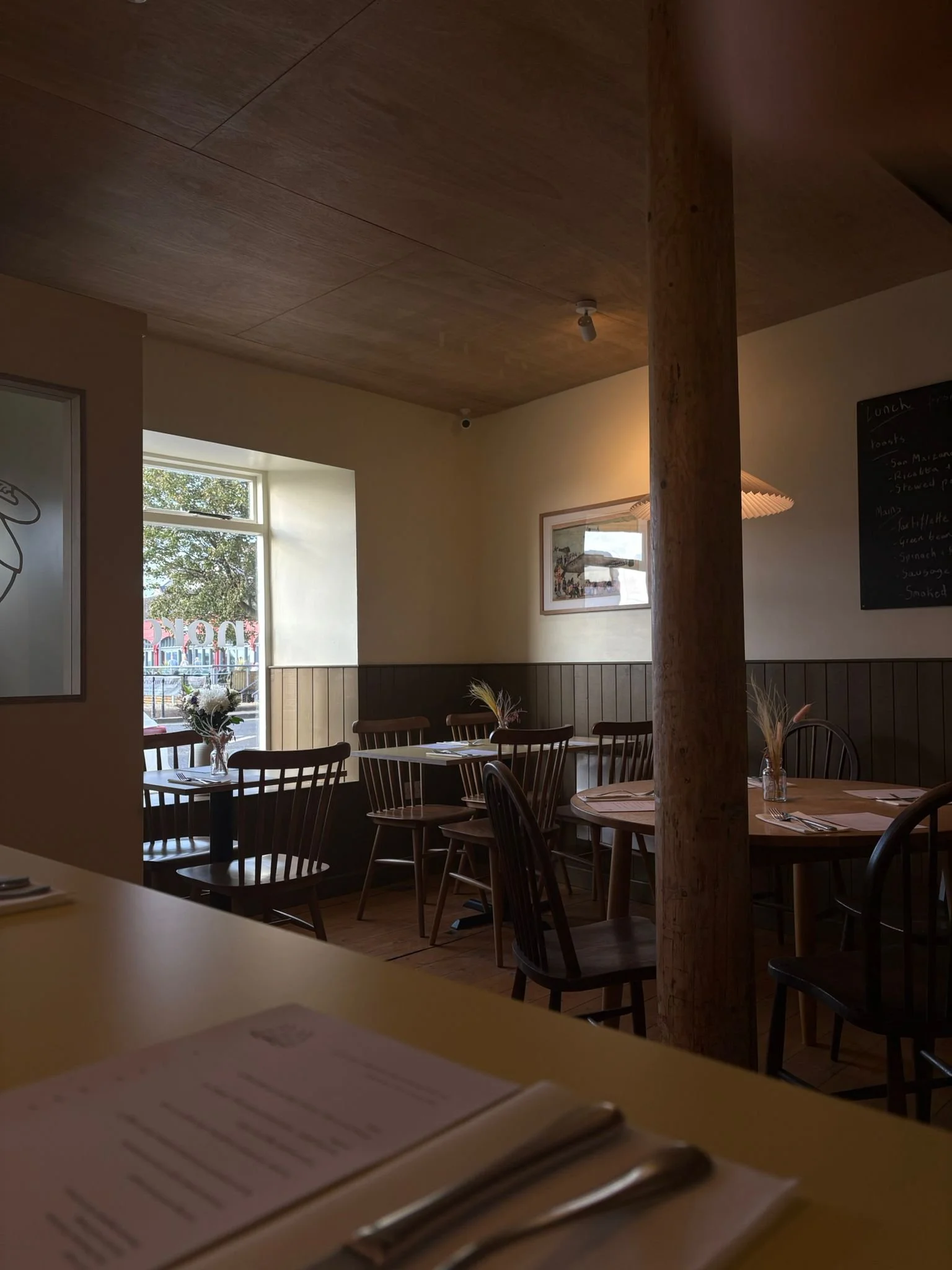 Interior of a cozy restaurant with wooden tables and chairs, a large window letting in natural light, and minimal decor including a blackboard menu and framed artwork.